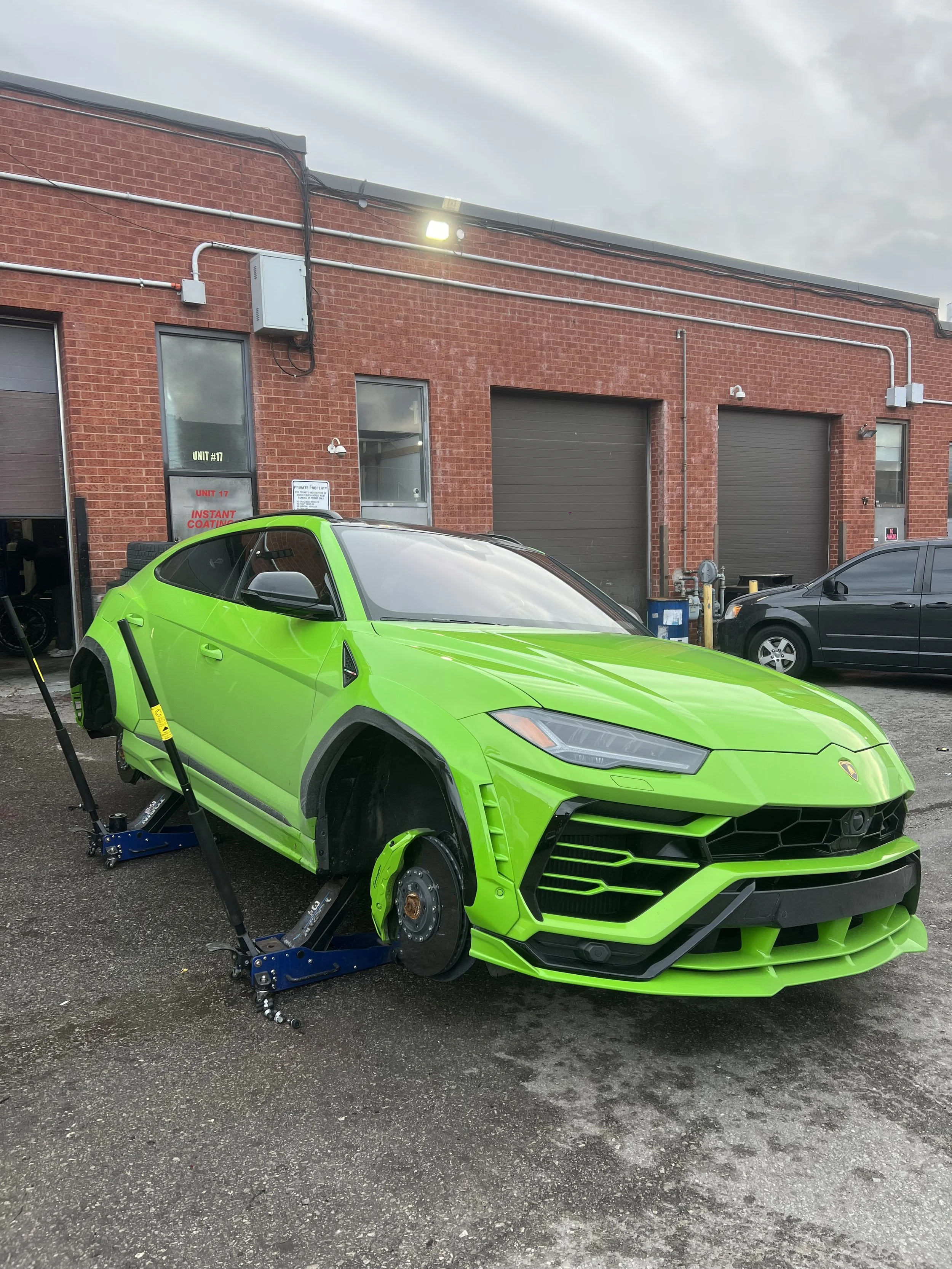 A bright green Lamborghini Urus undergoing repair, raised on a hydraulic lift with the front wheel removed, outside a red brick building.