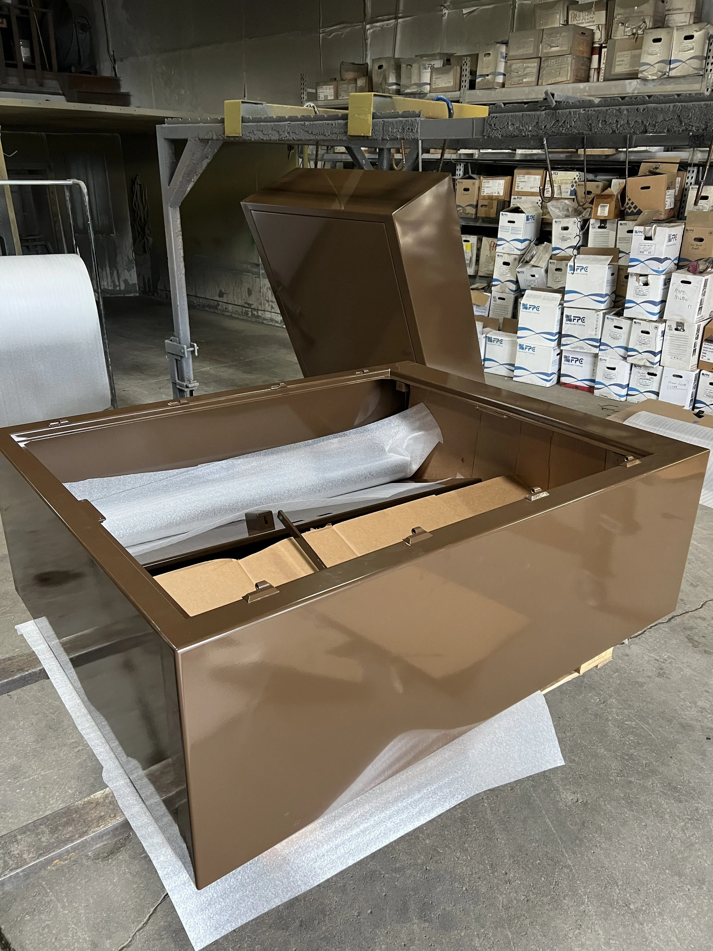 A large, rectangular, brown metallic freezer with an open lid, in a warehouse with shelves and boxes in the background.