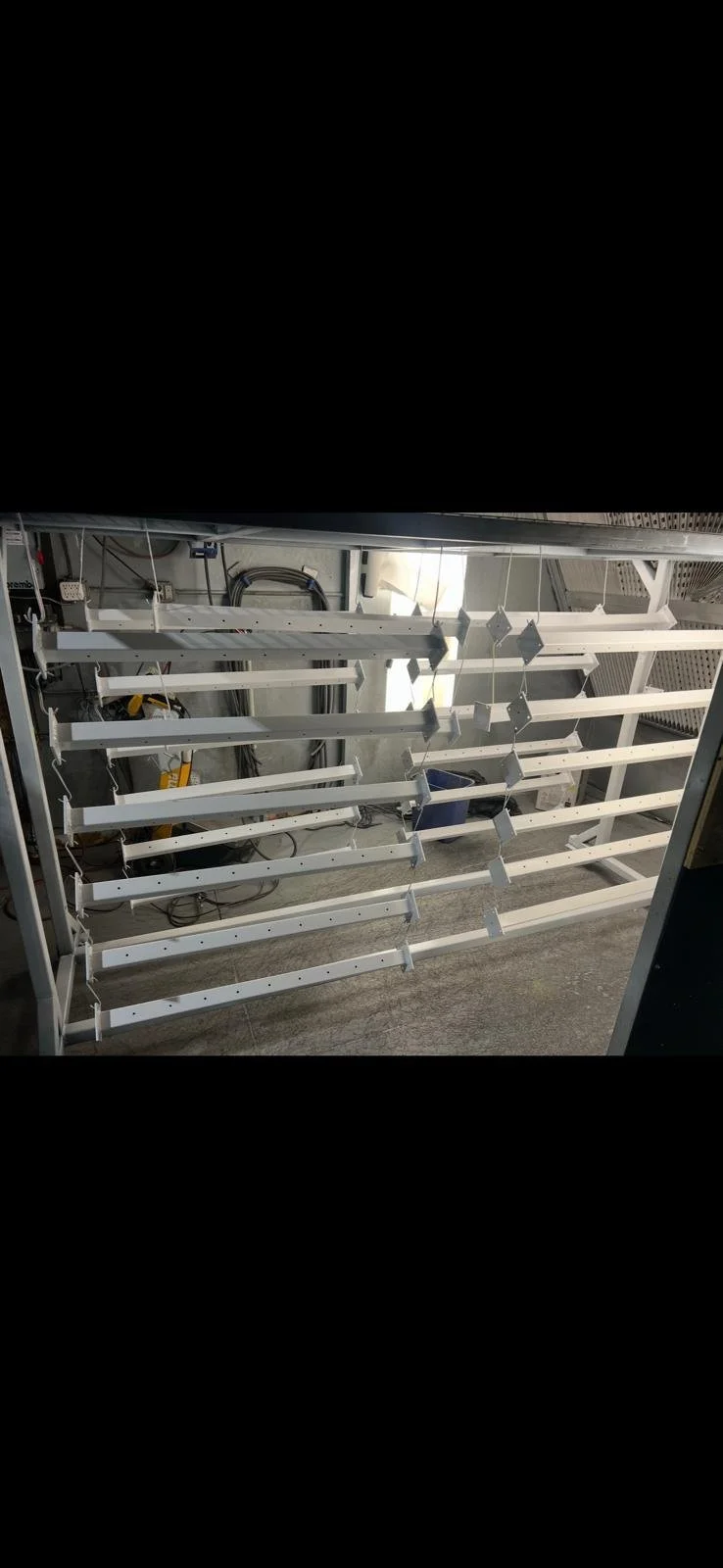 Empty white metal shelving units in a room with visible cables and equipment, possibly in a storage or maintenance area.