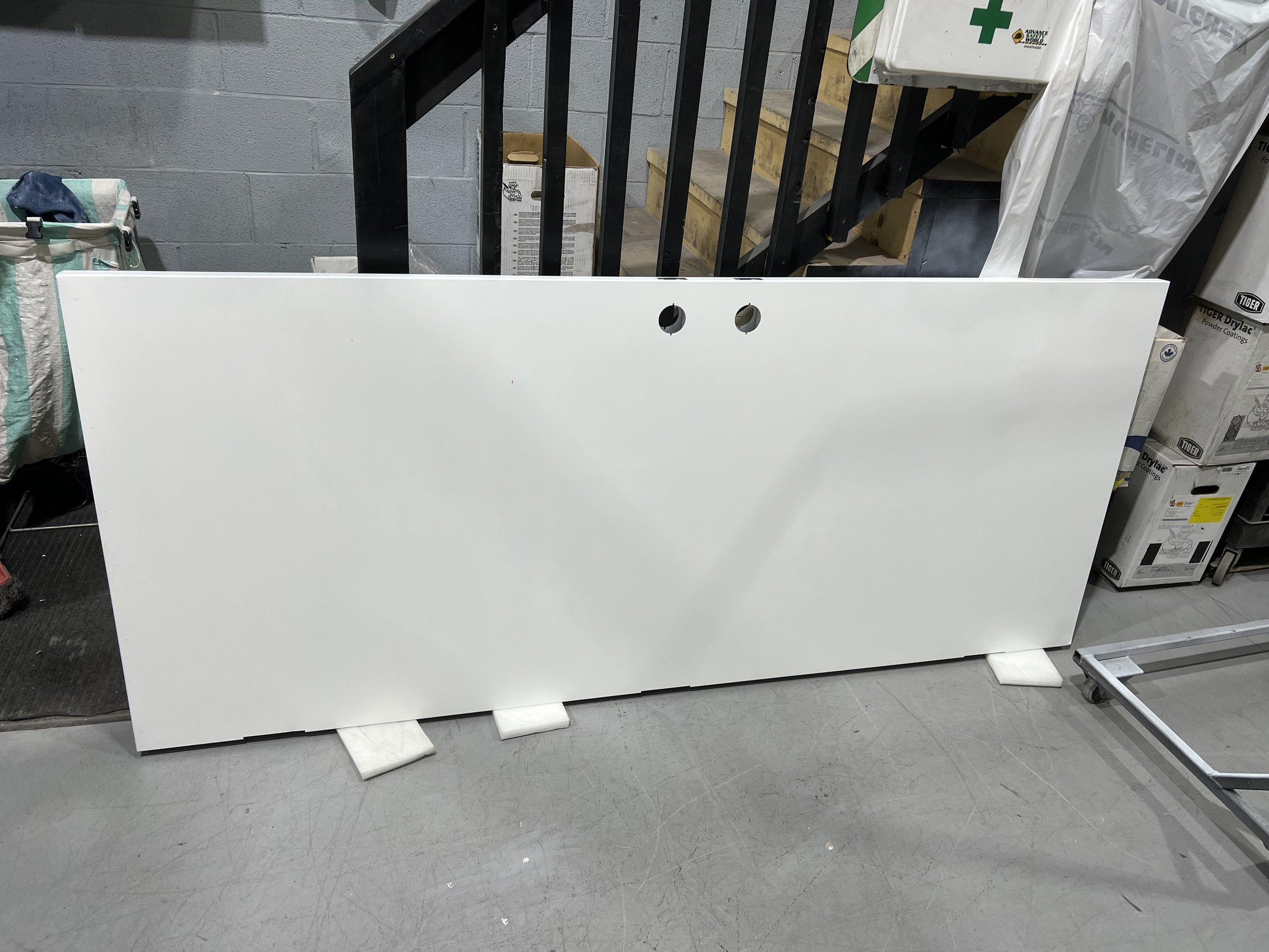 Large white rectangular panel resting on the floor, propped up with foam blocks, in a storage room with various boxes and equipment around.