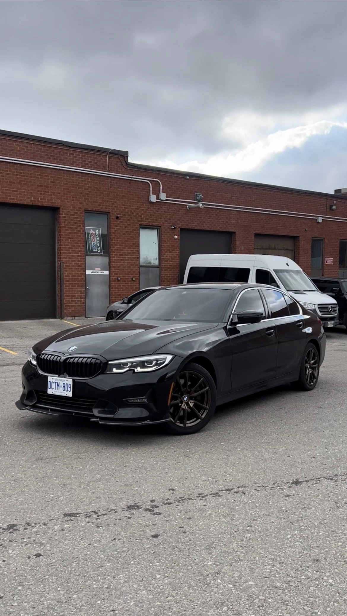 A black BMW sedan parked in a lot with a brick building and a white van in the background. Overcast sky. BRONZE RIMS