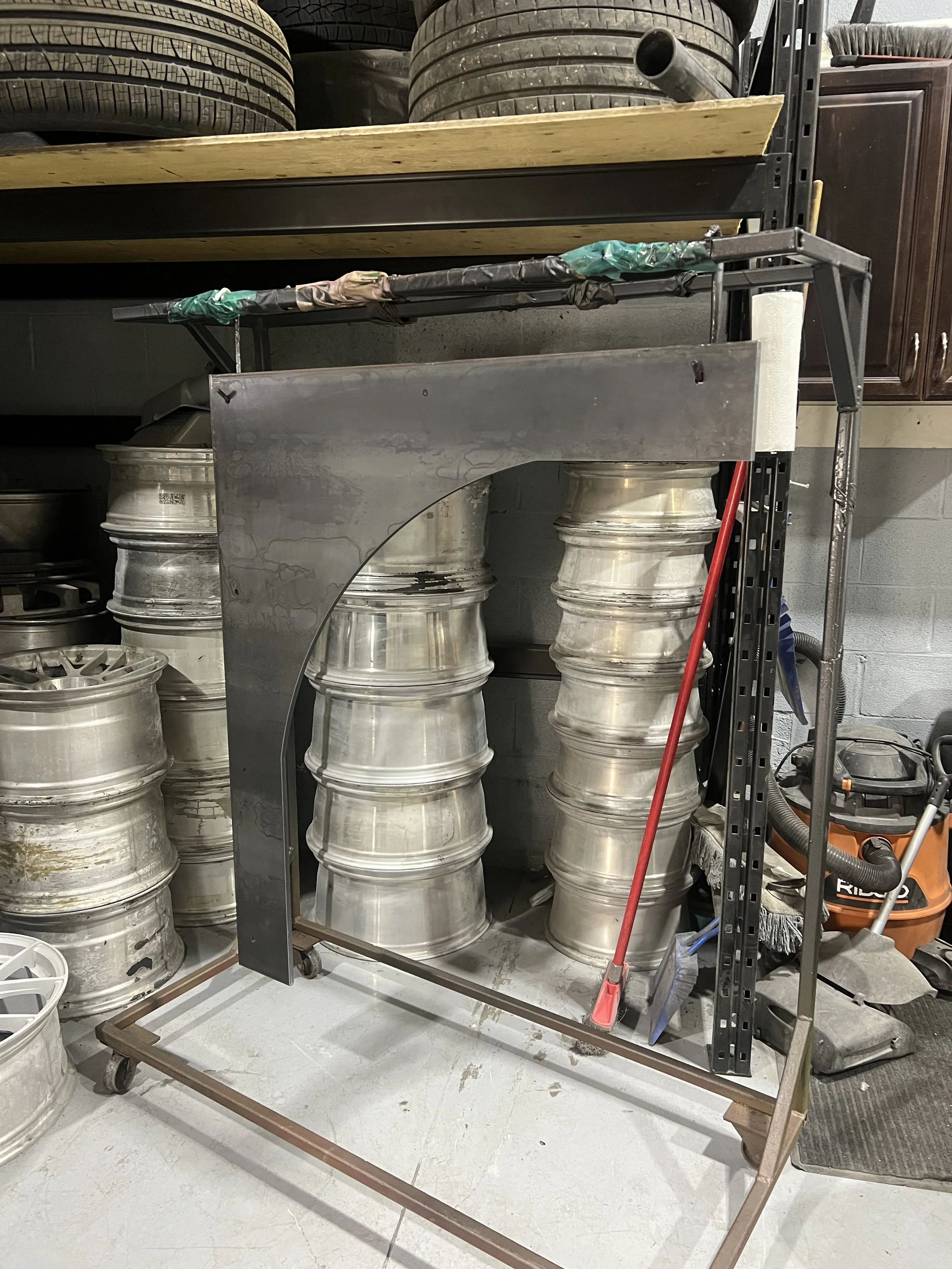 A metal sheet with a curved cutout leaning against a metal shelving unit. On the shelves above are car tires and other miscellaneous items. Various wheel rims are stacked on the floor behind the sheet, and cleaning equipment like a broom and vacuum c