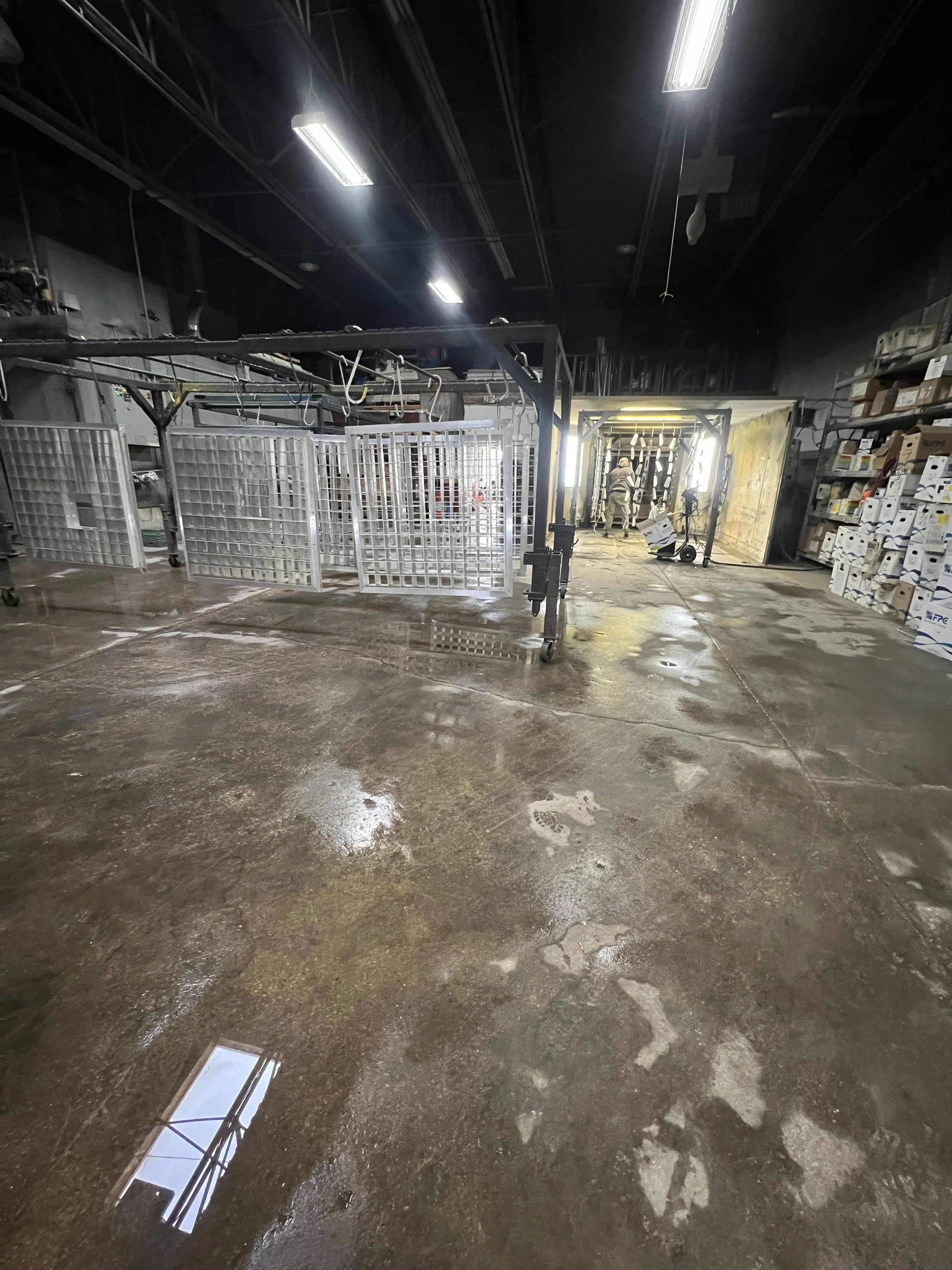 Indoor construction or storage area with wet, stained concrete floor, metal carts and frames, shelves with boxes, and a worker in protective gear near a ladder and bright light at the far end.