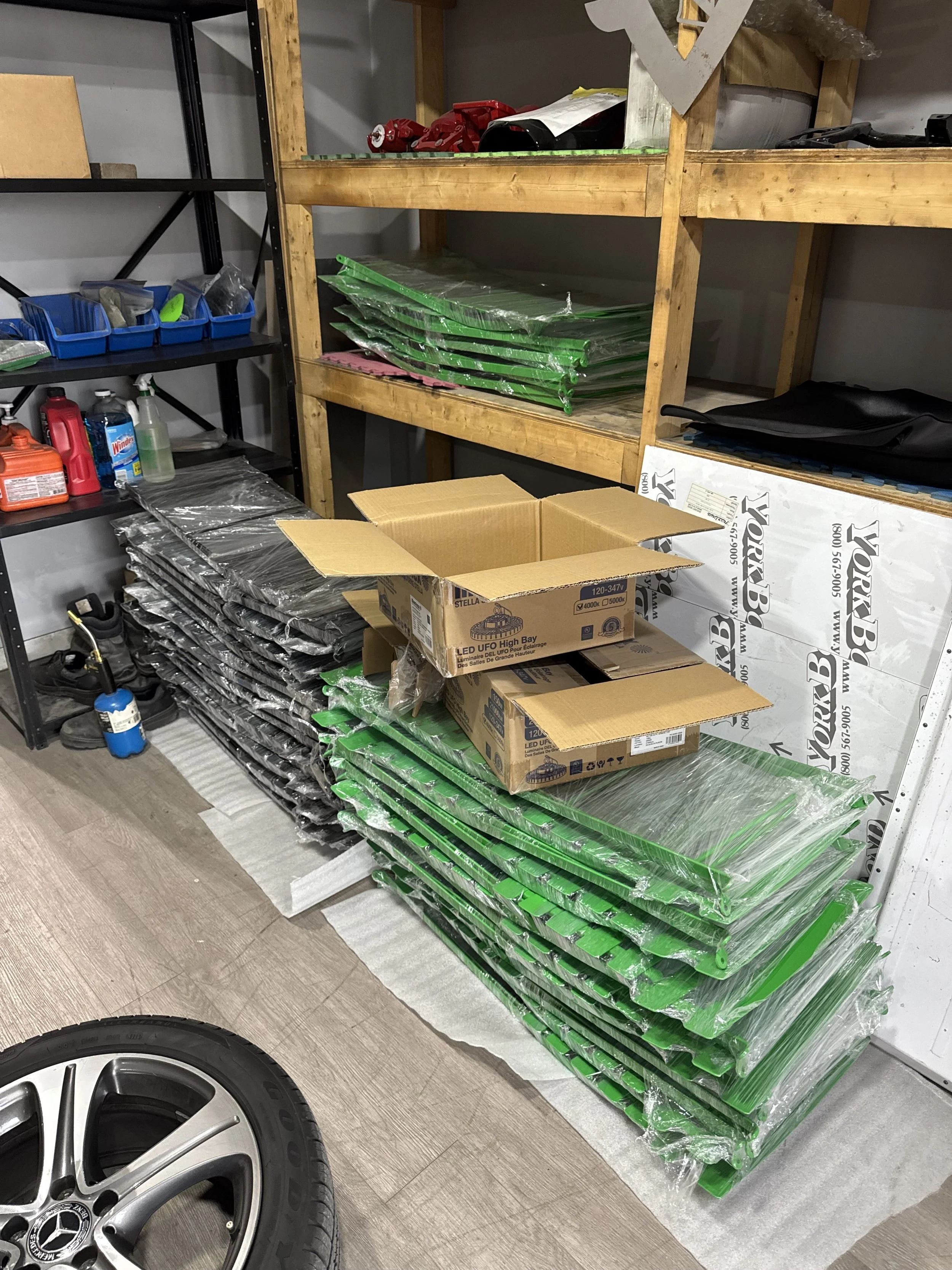 Shelving unit with various storage items, green plastic tents stacked on the floor, open cardboard boxes, and a car wheel in the foreground.