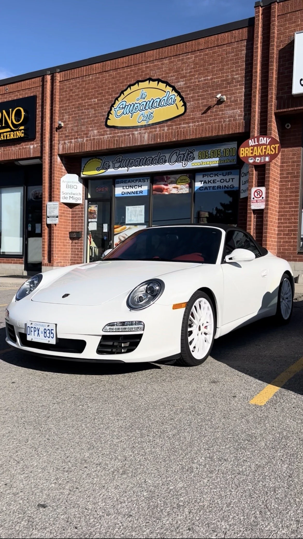 A white Porsche convertible car parked in front of La Ecuadoreña Café, a brick building with signage advertising breakfast, lunch, dinner, and catering services.