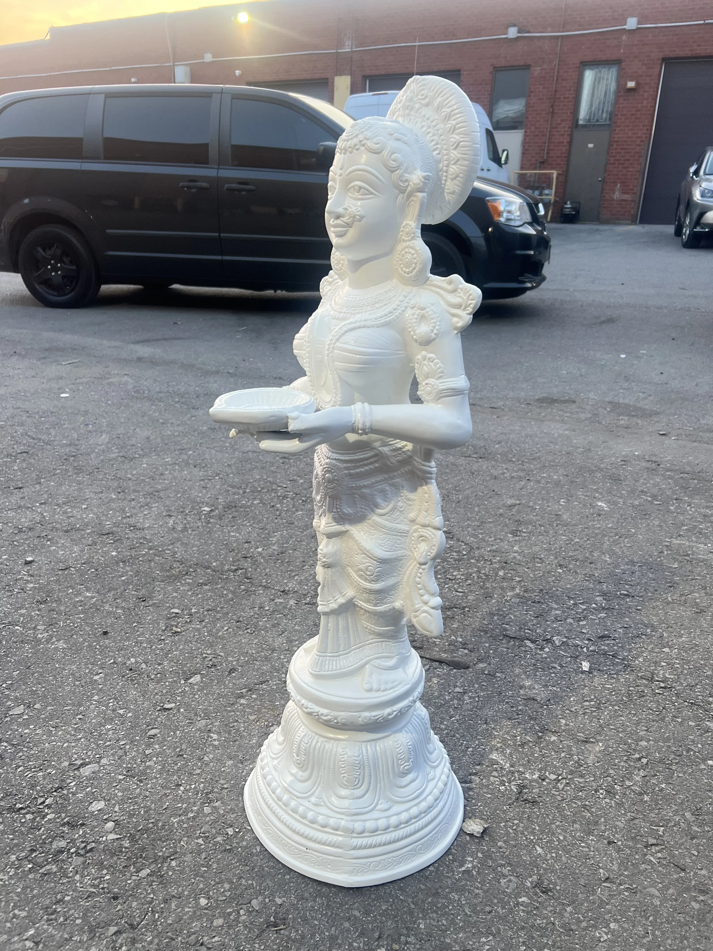 White carved statue of a woman in traditional attire holding a dish, standing outdoors on asphalt with vehicles and brick buildings in the background.
