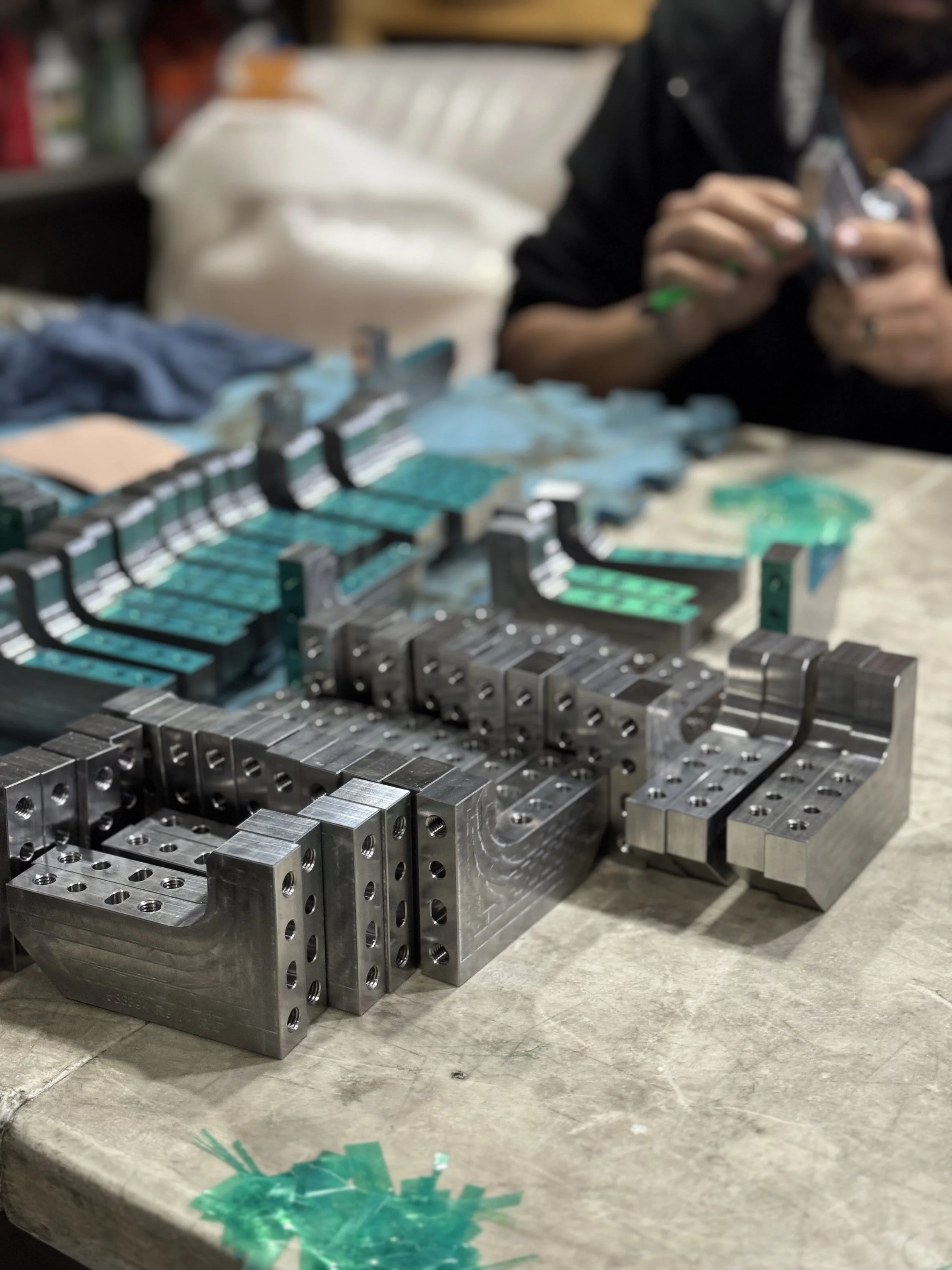 Close-up of metal machine parts with wires on a workbench, with a person in the background using a tool.