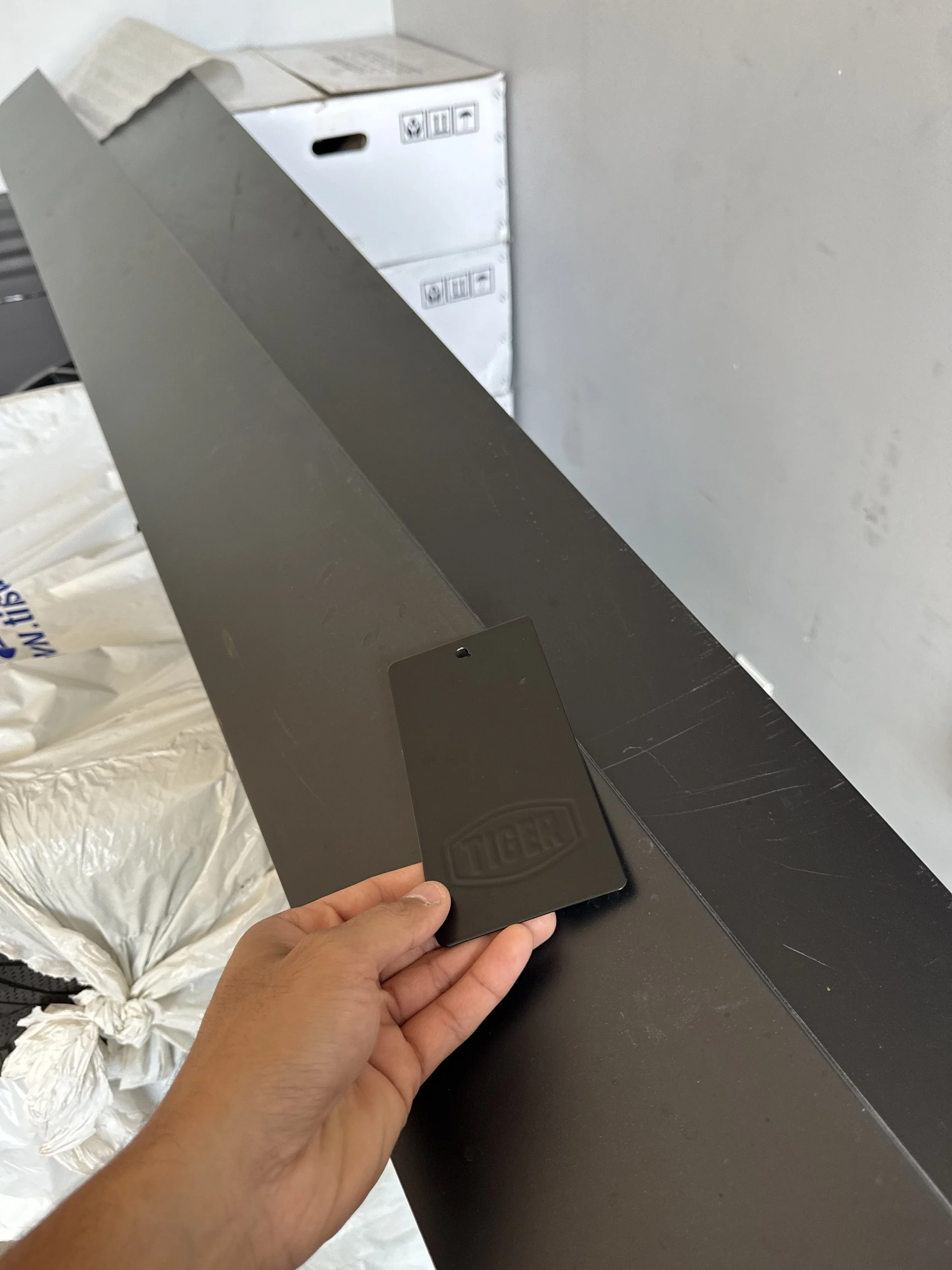 A person is holding a black metal object with the brand name 'TIGER' embossed on it, resting on a large flat black surface, with boxes in the background.