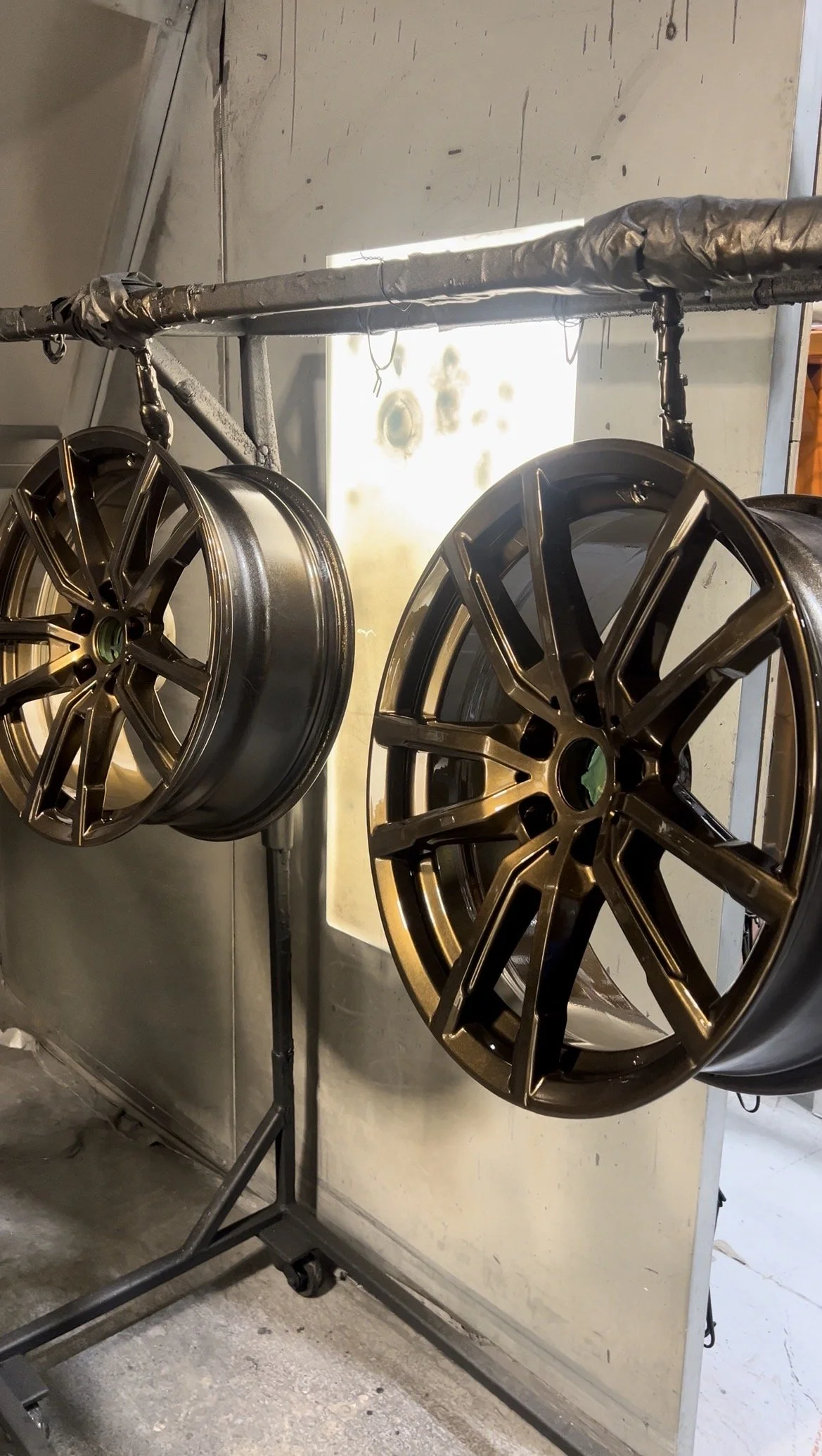 Two black automotive wheels hanging from a ceiling, possibly in a workshop, with a wall, a small window, and a metallic surface in the background.