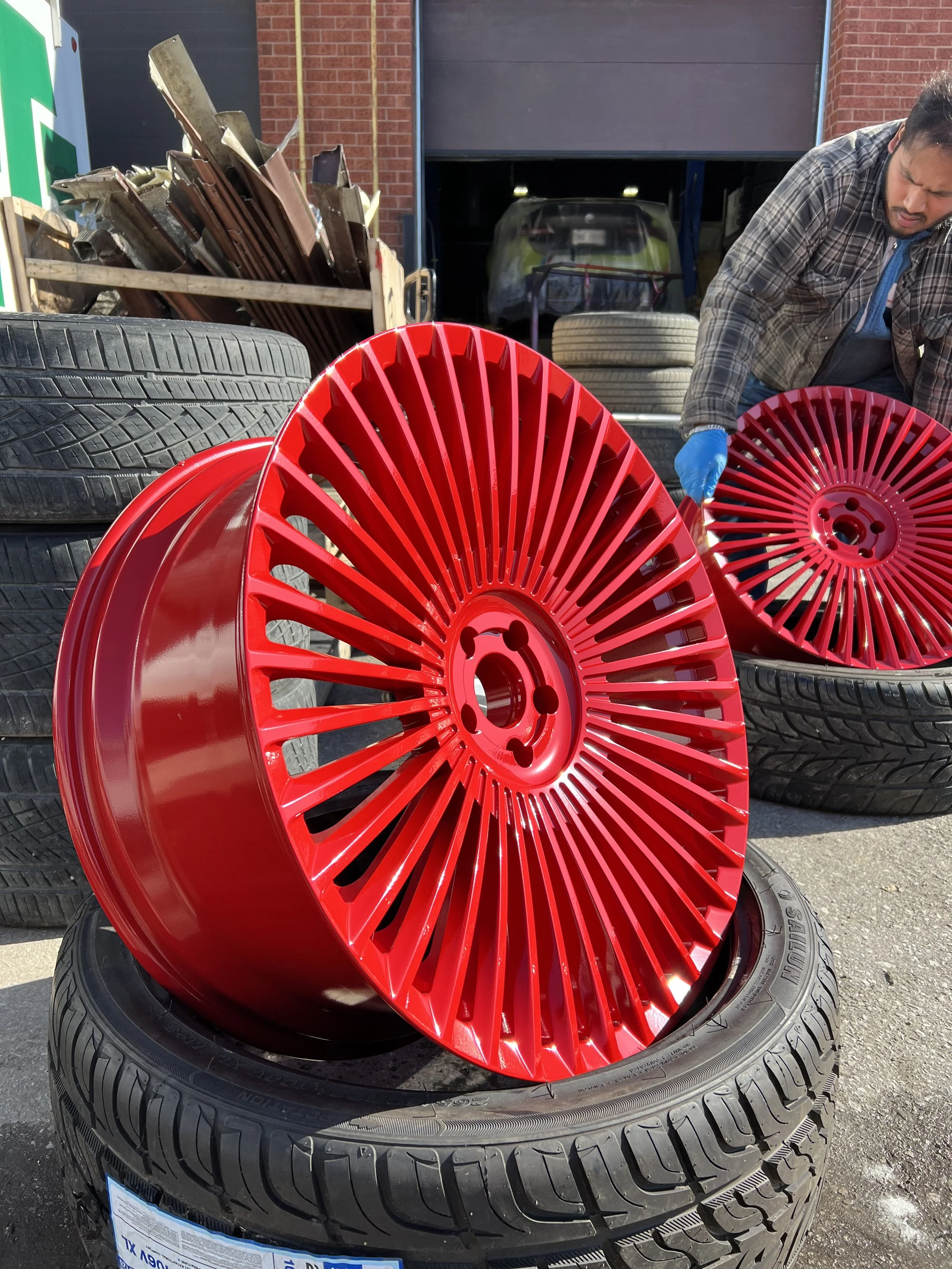 Red car wheel rims stacked on tires outside auto shop with man working on a wheel in the background.