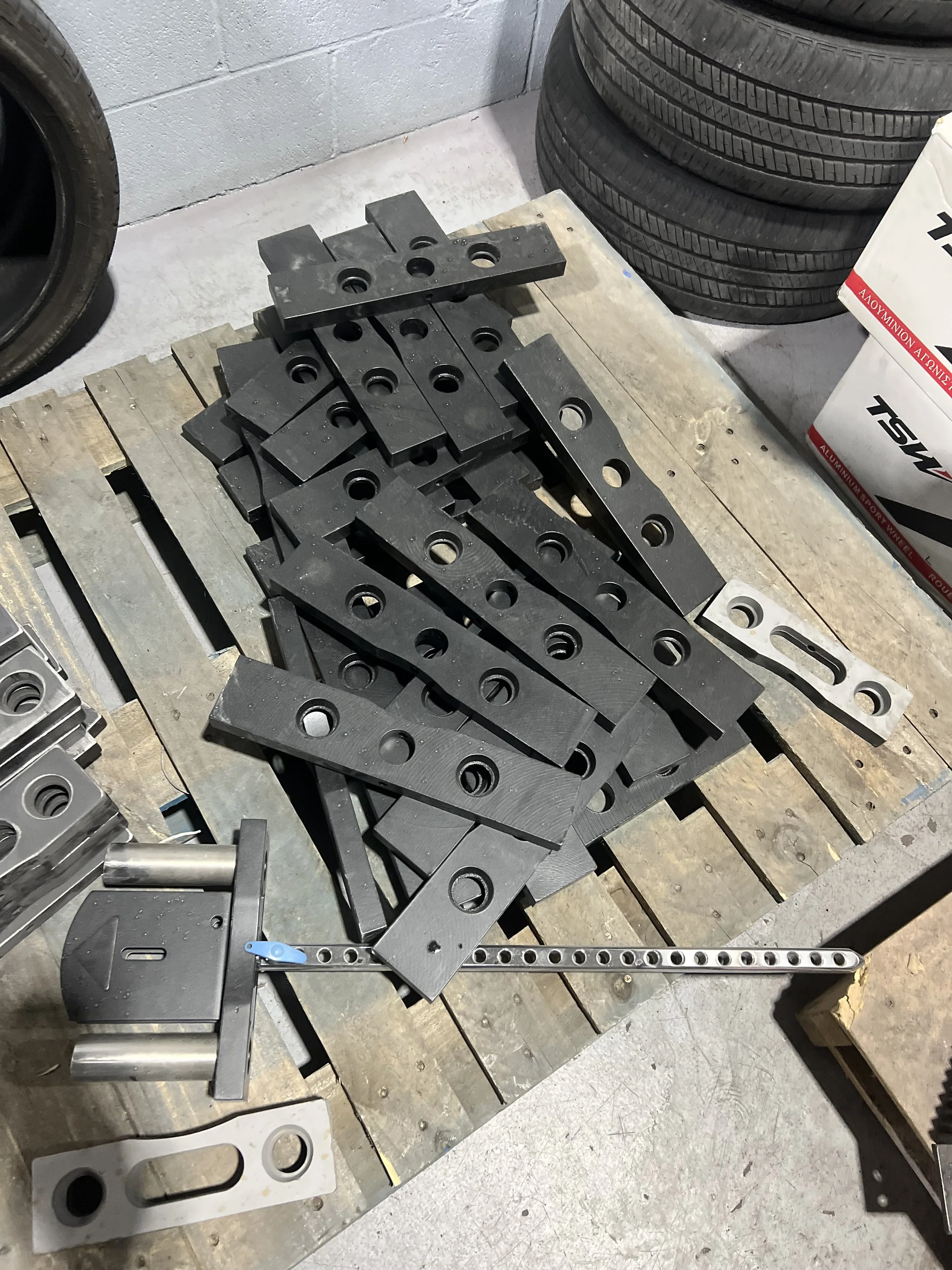 Collection of black metal plates with holes, arranged on a wooden pallet in a workshop or garage setting.