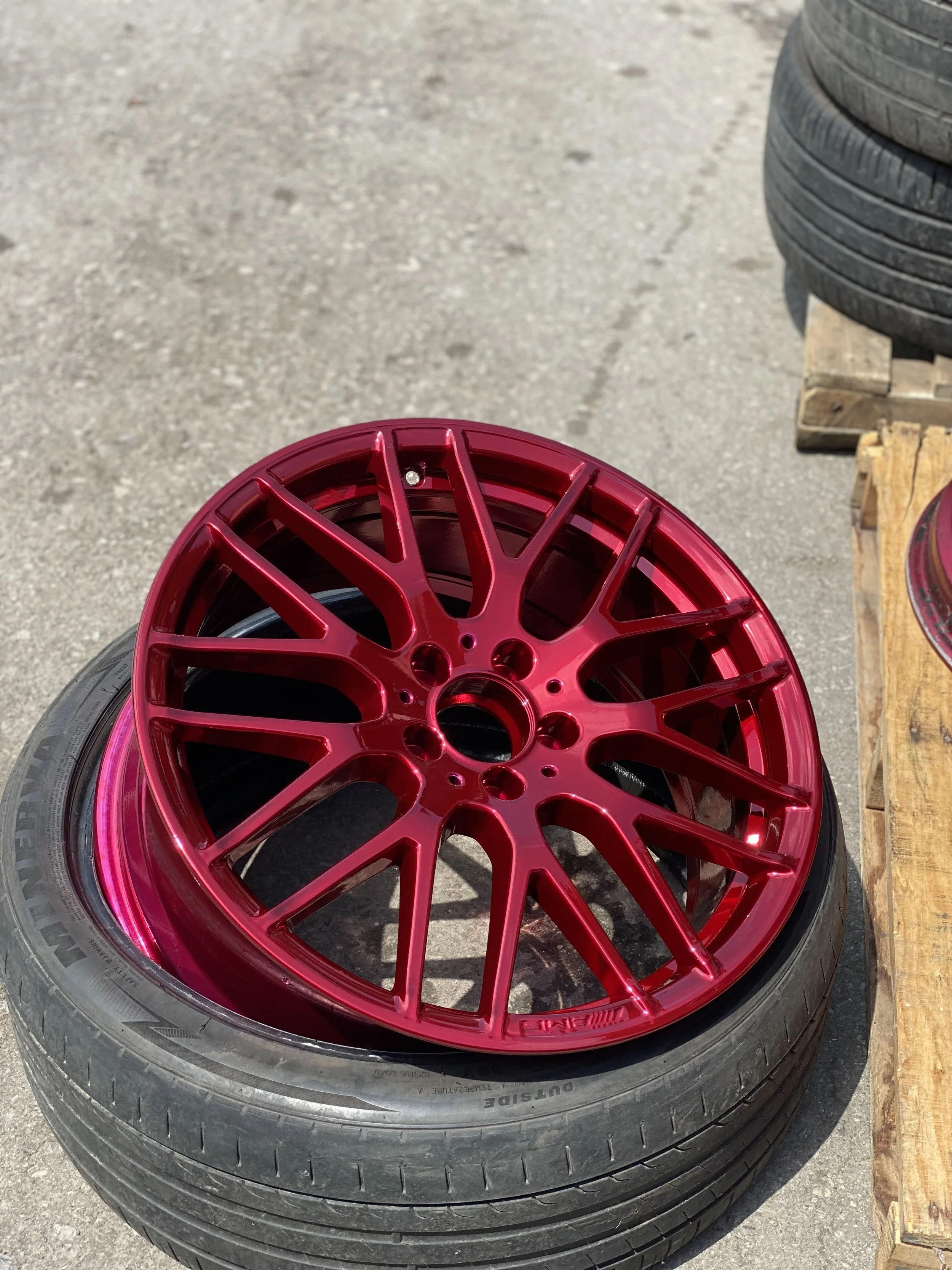 A red aftermarket car wheel rim resting on a tire, outdoors on a concrete surface, with stacks of other tires nearby.