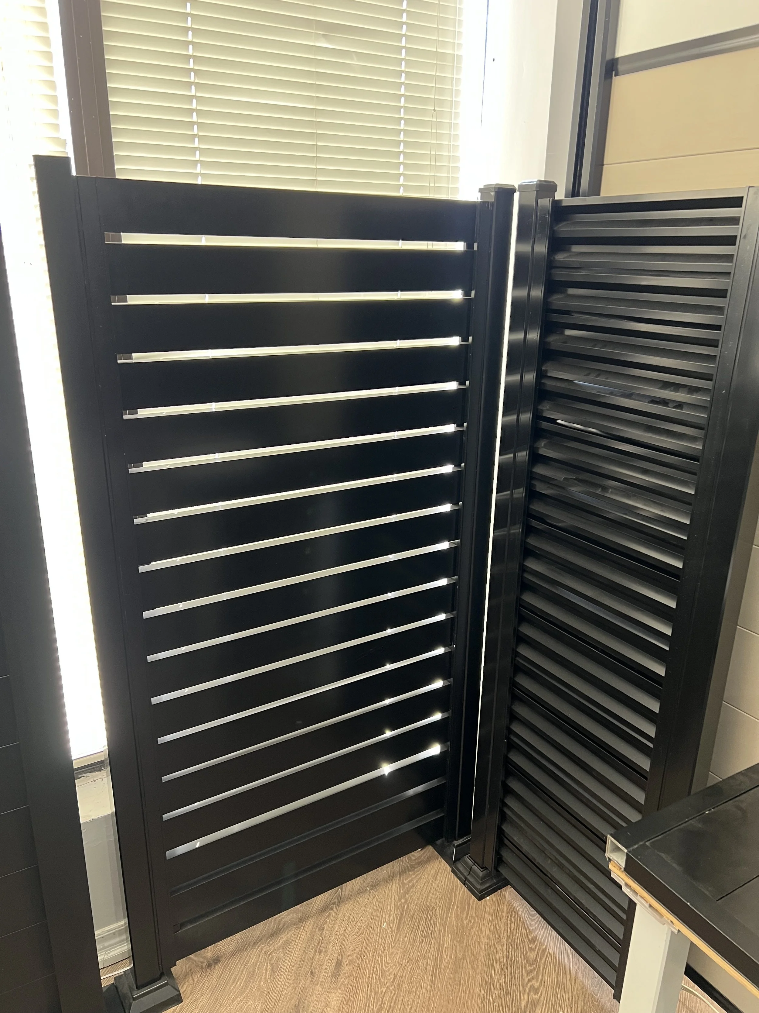 A black louvered door with horizontal slats, open at the top, standing indoors near a window with closed blinds, next to a second matching black louvered door.
