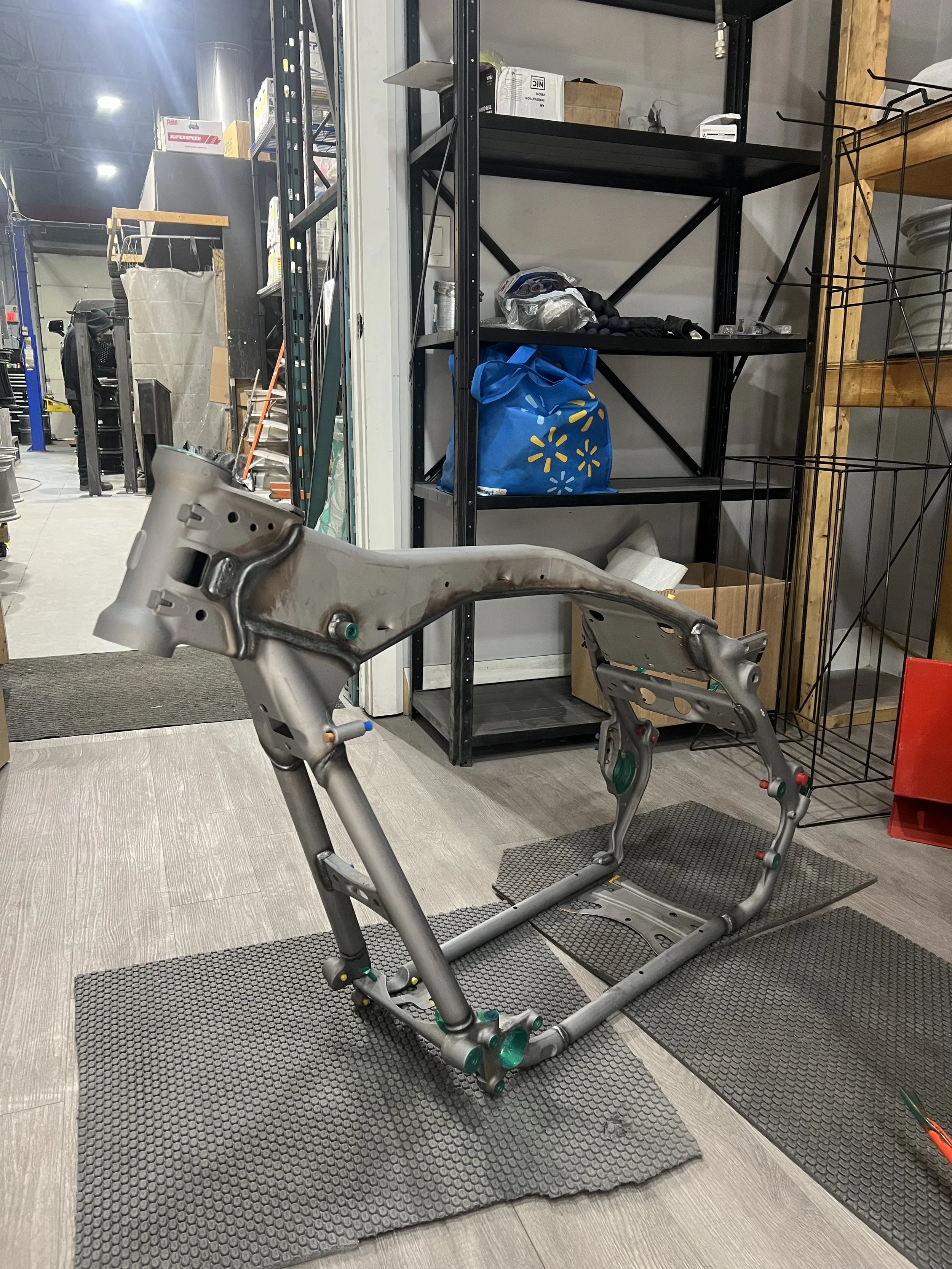 Metal framework of a bicycle or motorcycle in a workshop or warehouse setting, with shelves and boxes in the background.