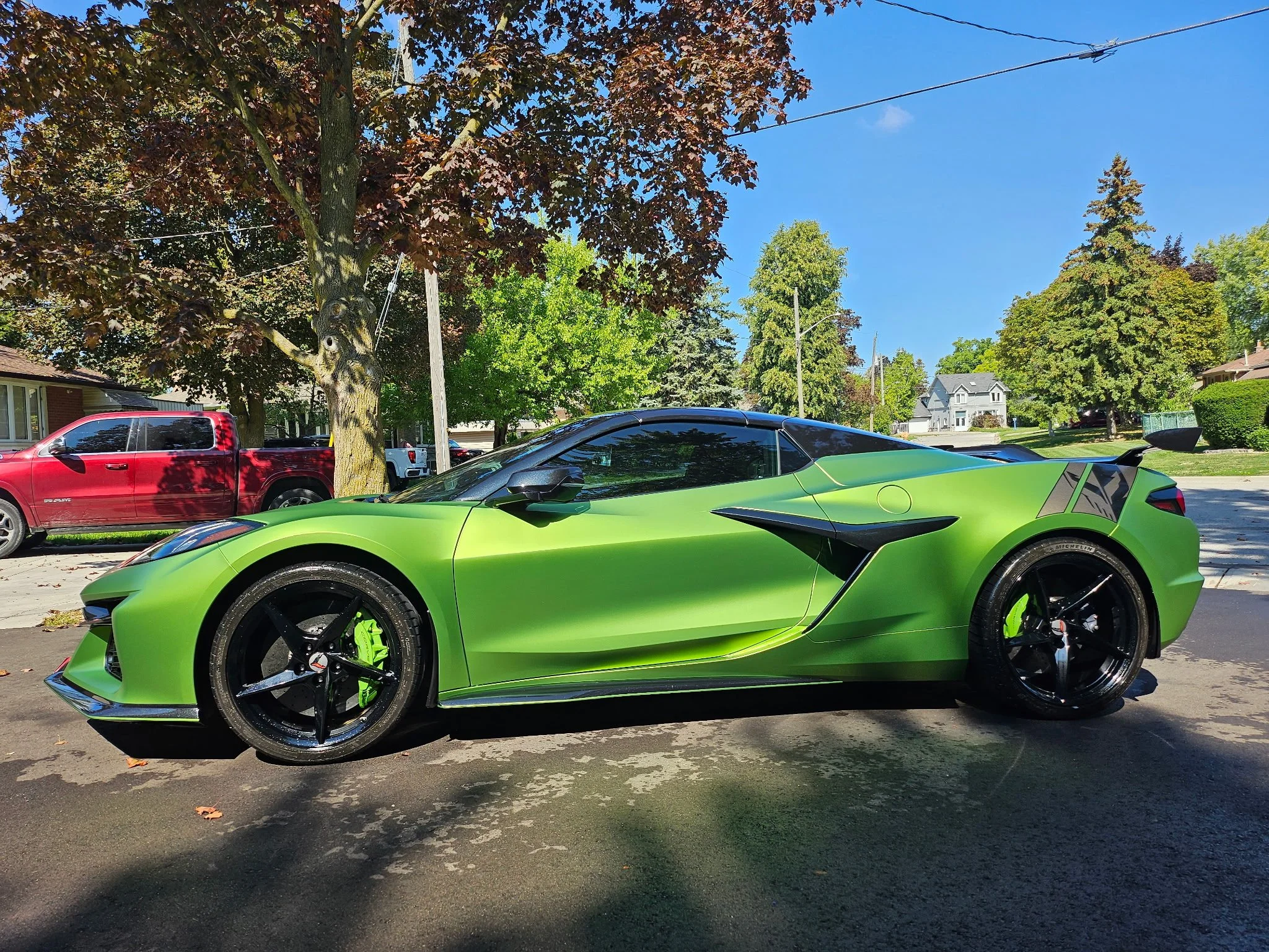 A bright green sports car parked on a street with trees and houses in the background. A red pickup truck is visible to the left.