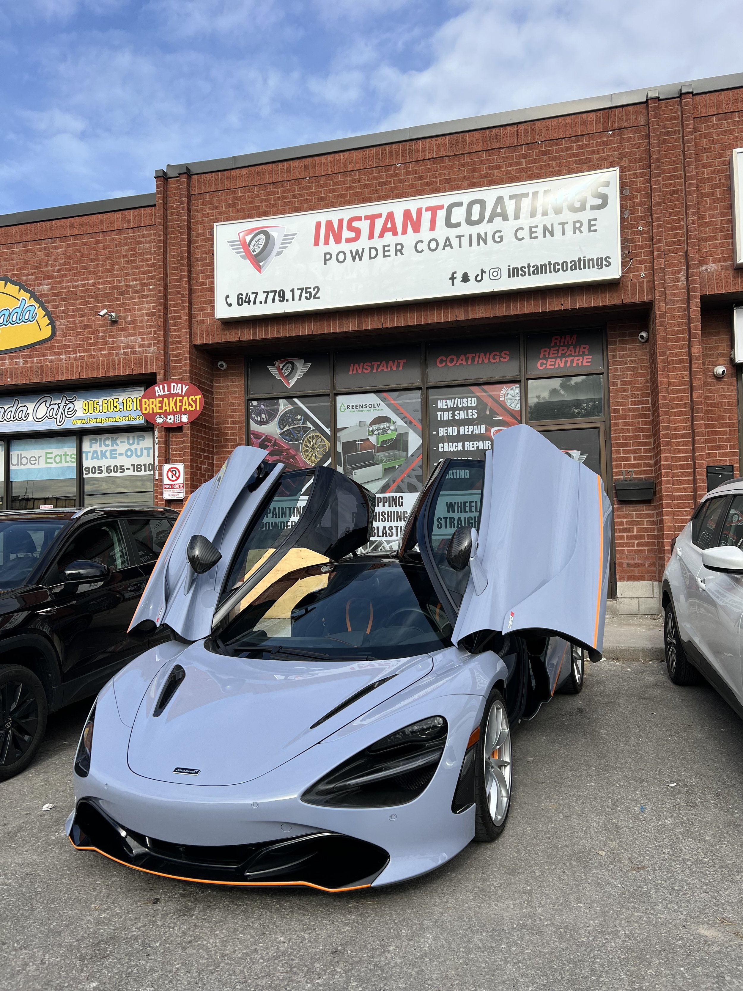 A silver sports car with gullwing doors open parked in front of a brick building with a sign that reads 'Instant Coatings Powder Coating Centre' and a storefront with additional signs for tire repairs and services.
