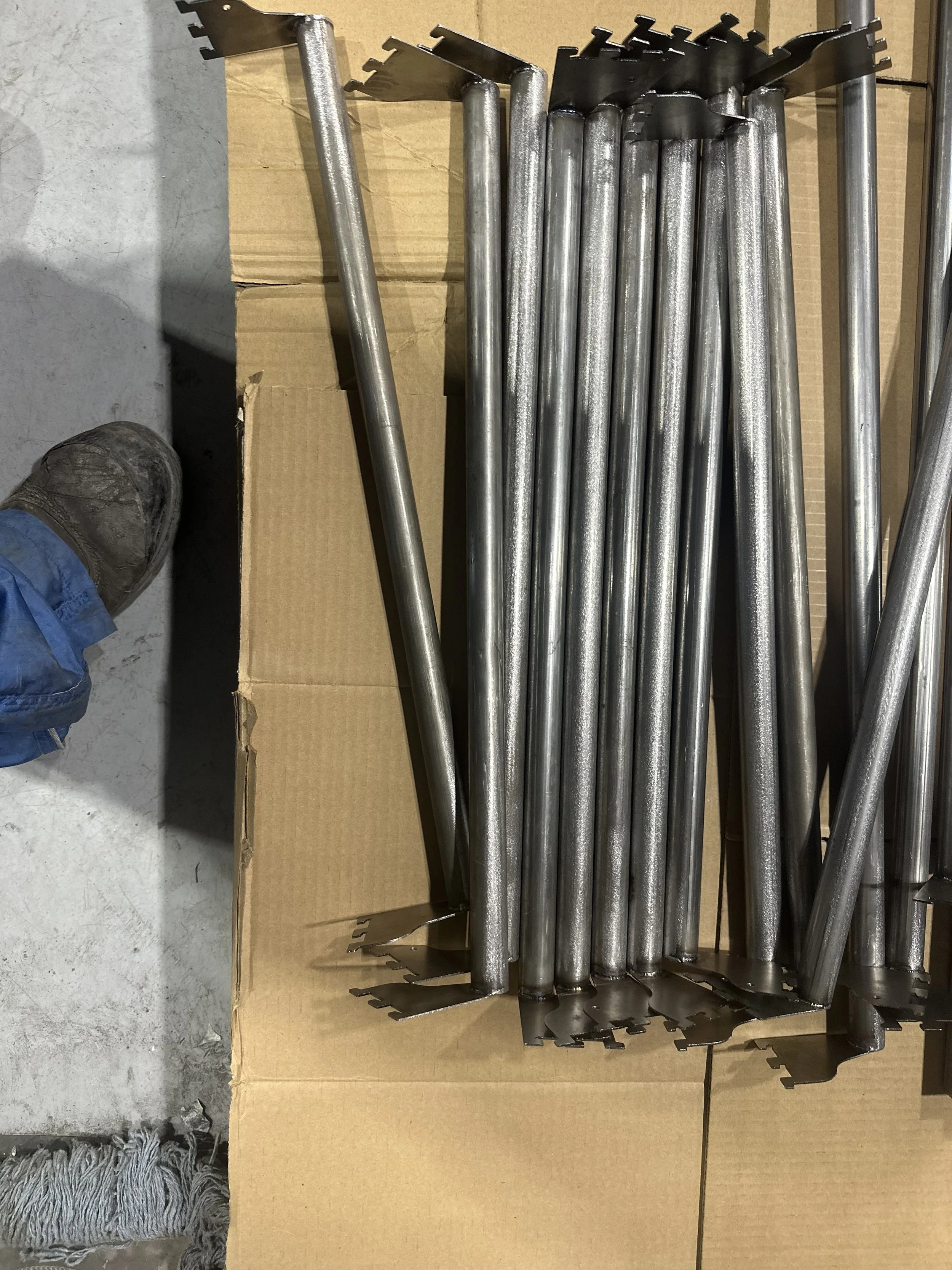 Metal stakes stacked in a cardboard box, with a person's foot in a work boot visible on the left side.