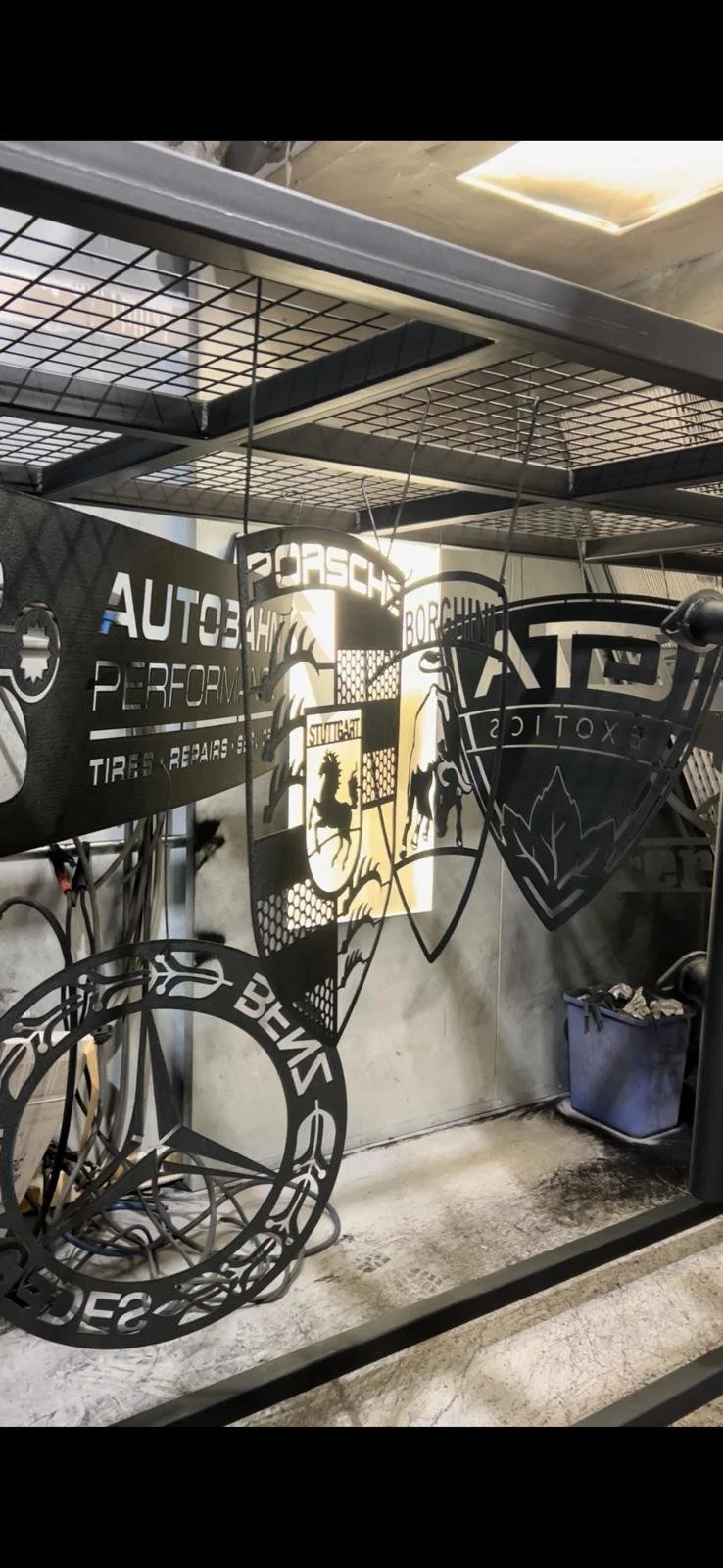 Metal car brand logos and signs hanging on a wire rack in a garage or workshop setting.