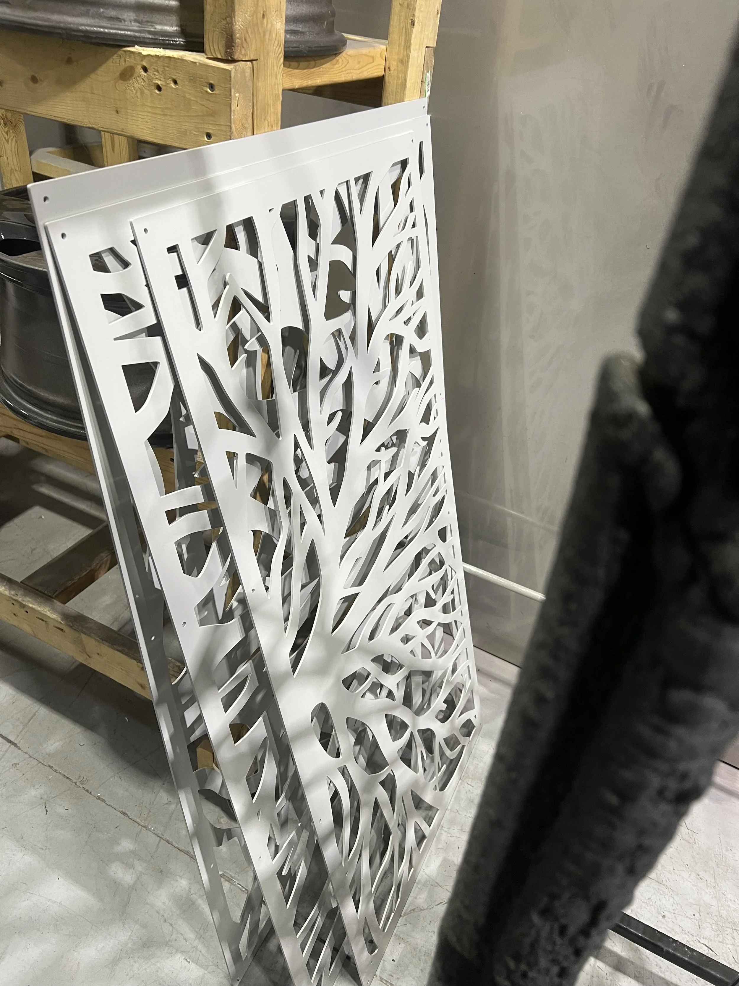 White metal decorative panel with a tree and leaf design leaning against a wooden shelf in a workshop or storage area.