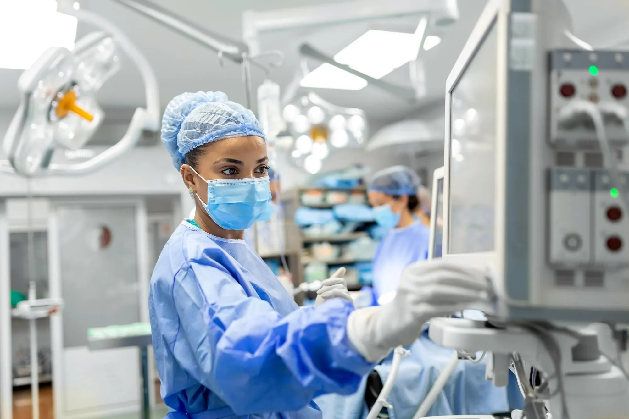 A female medical professional in a hospital operating room, wearing a blue surgical gown, mask, cap, and gloves, working with medical equipment.