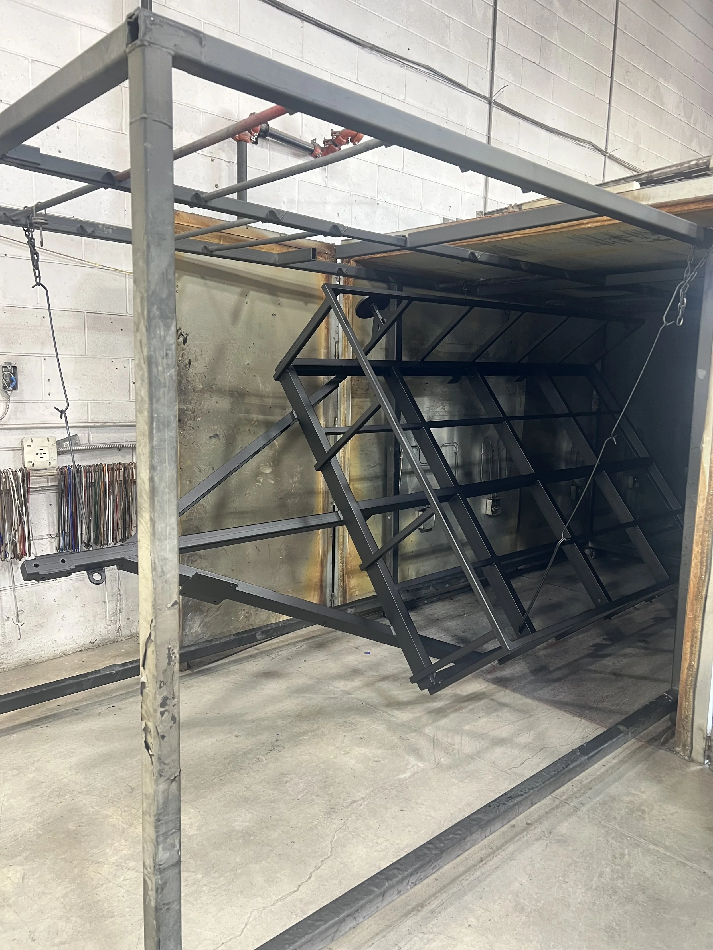 Metal frame structure, possibly part of a scaffolding or manufacturing bed, with black metal components and a support grid. The background includes a concrete wall with some piping and electrical outlets.