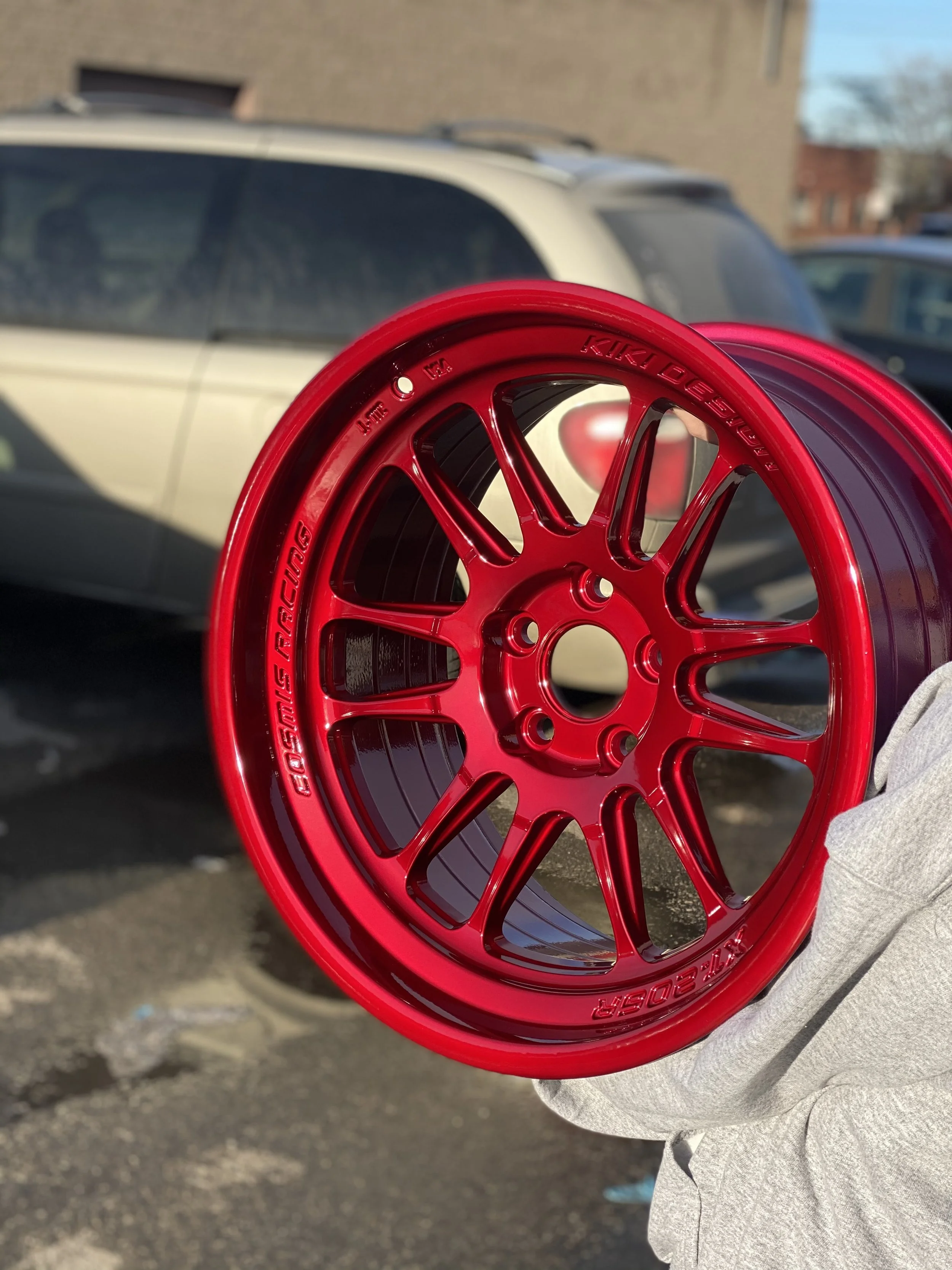 A shiny red car wheel rim being held by a person's hand in front of parked cars and a building.