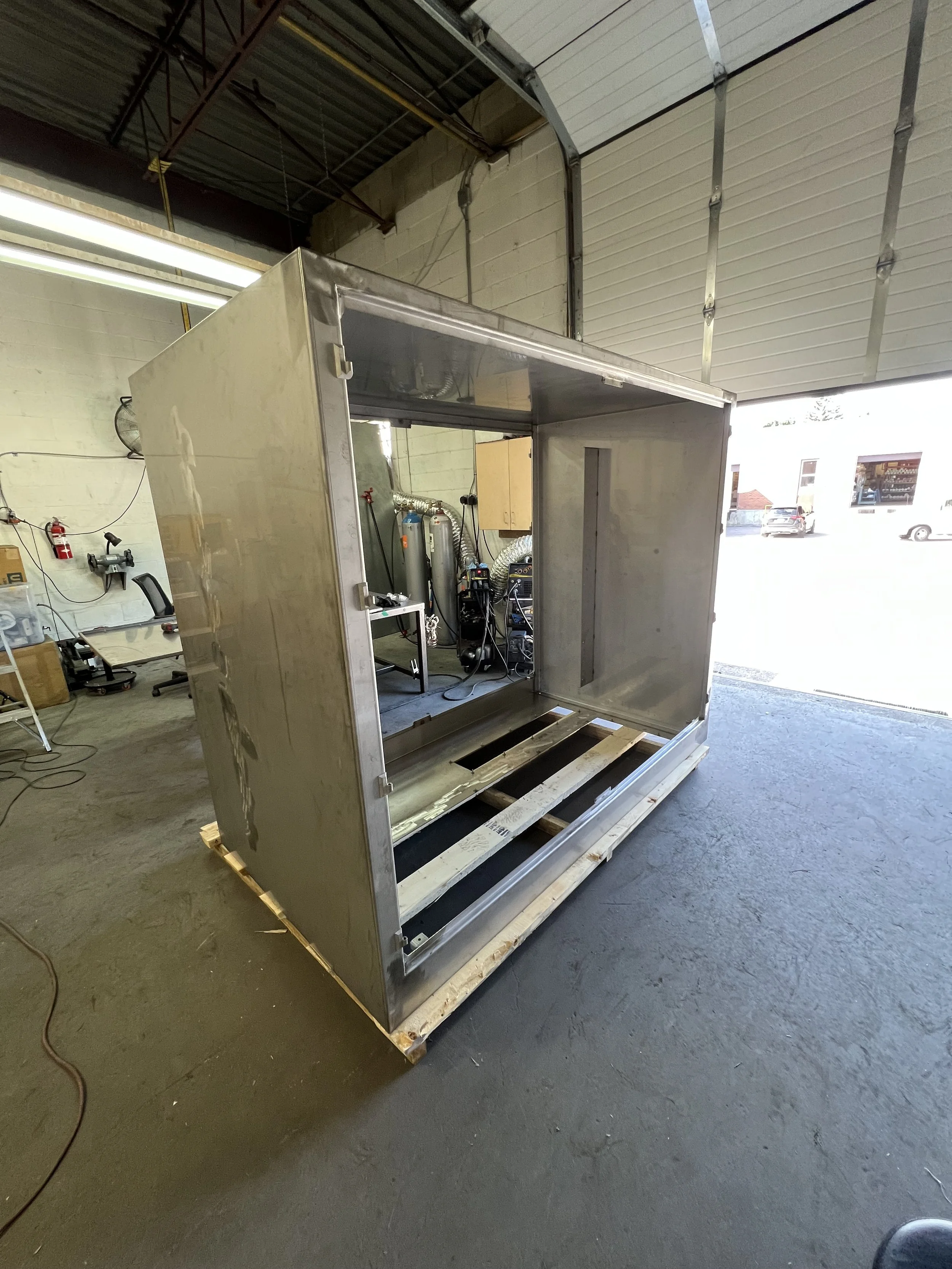 Unfinished stainless steel enclosure on a wooden pallet inside a garage or workshop, with tools and equipment in the background.