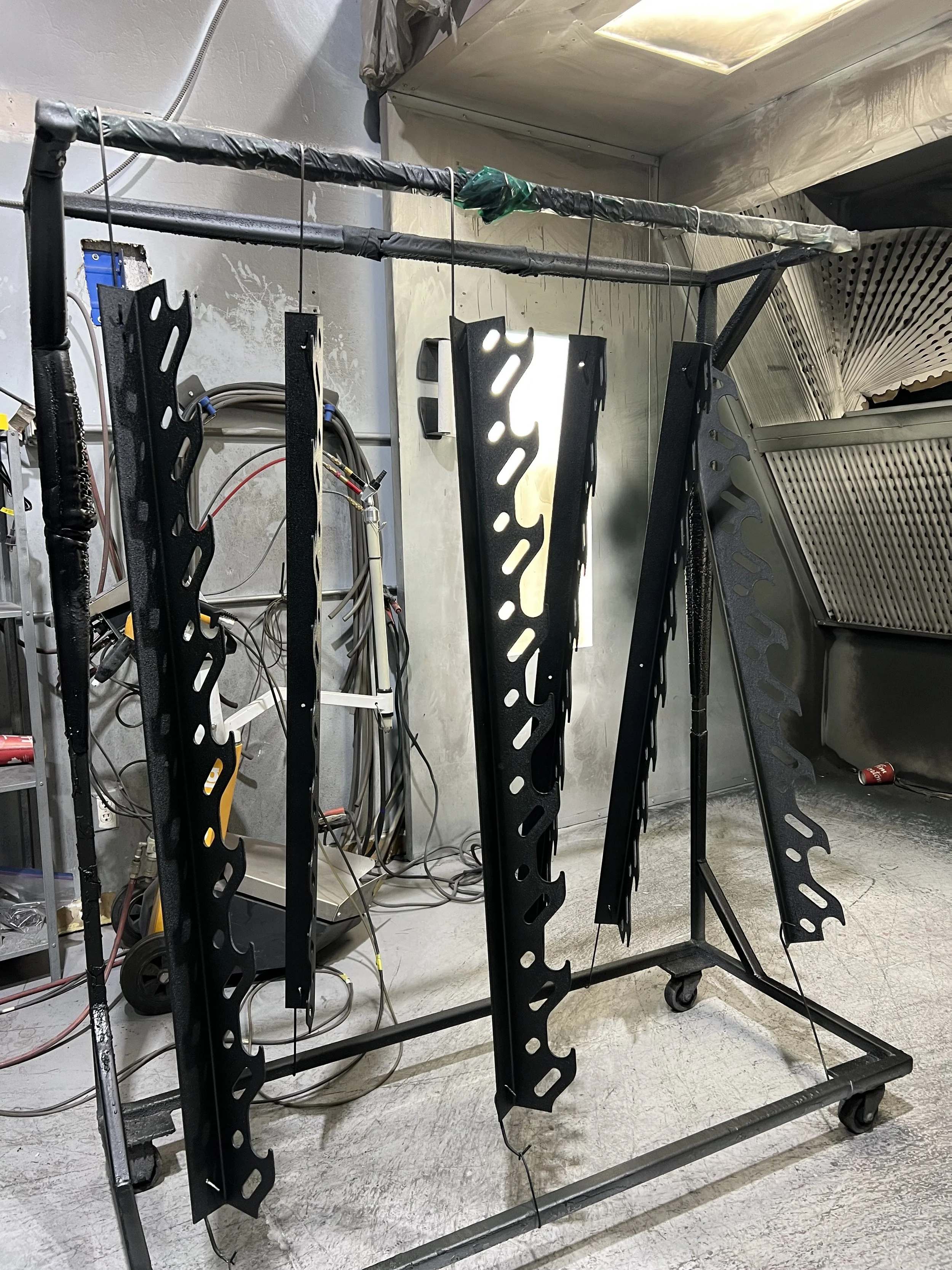 Metal framework with black painted parts hanging on wires in a workshop or manufacturing space.