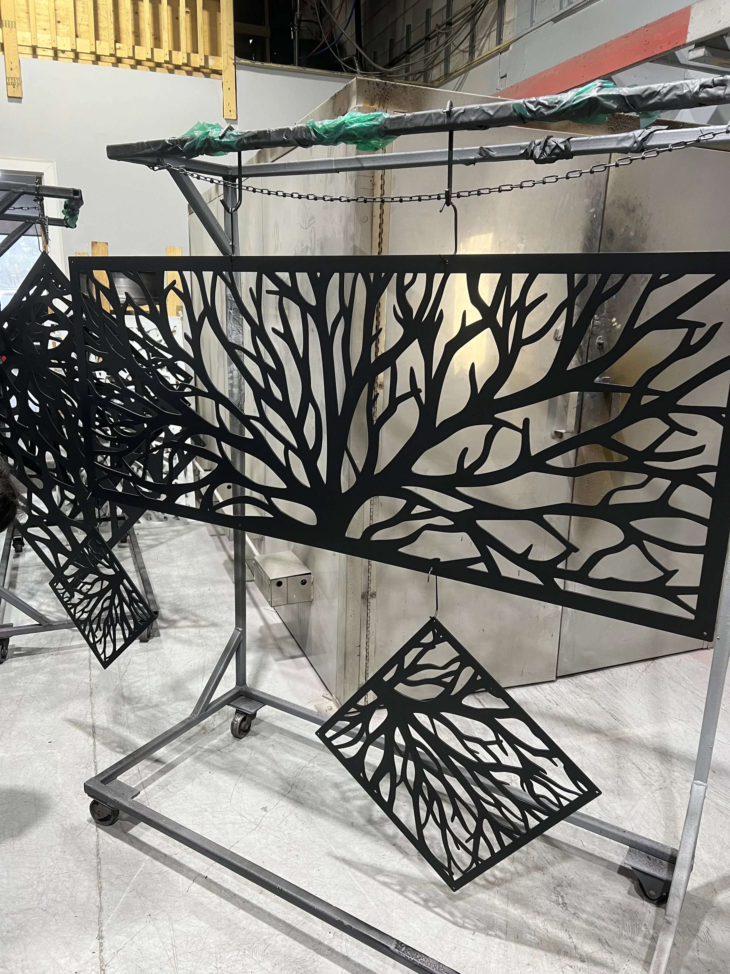 Black metal decorative panels with tree branch cut-out design hanging on a metal frame in a workshop.