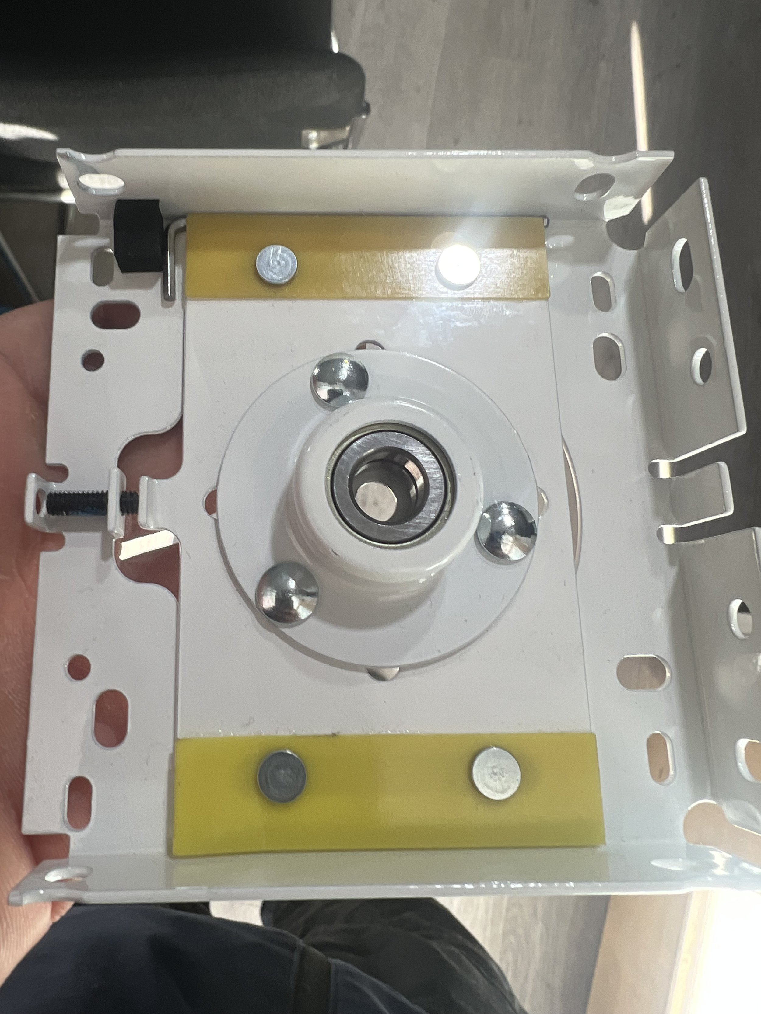 Open electrical box with metal mounting bracket and central bearing, yellow spacers, and visible screws.