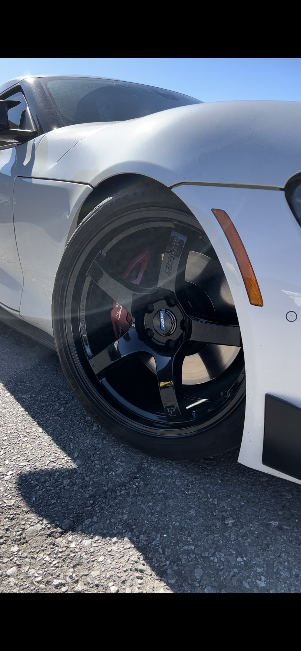 Close-up of a white sports car's front left side, showing black alloy wheels, red brake calipers, and a part of the headlight, on an outdoor gravel surface against a blue sky.
