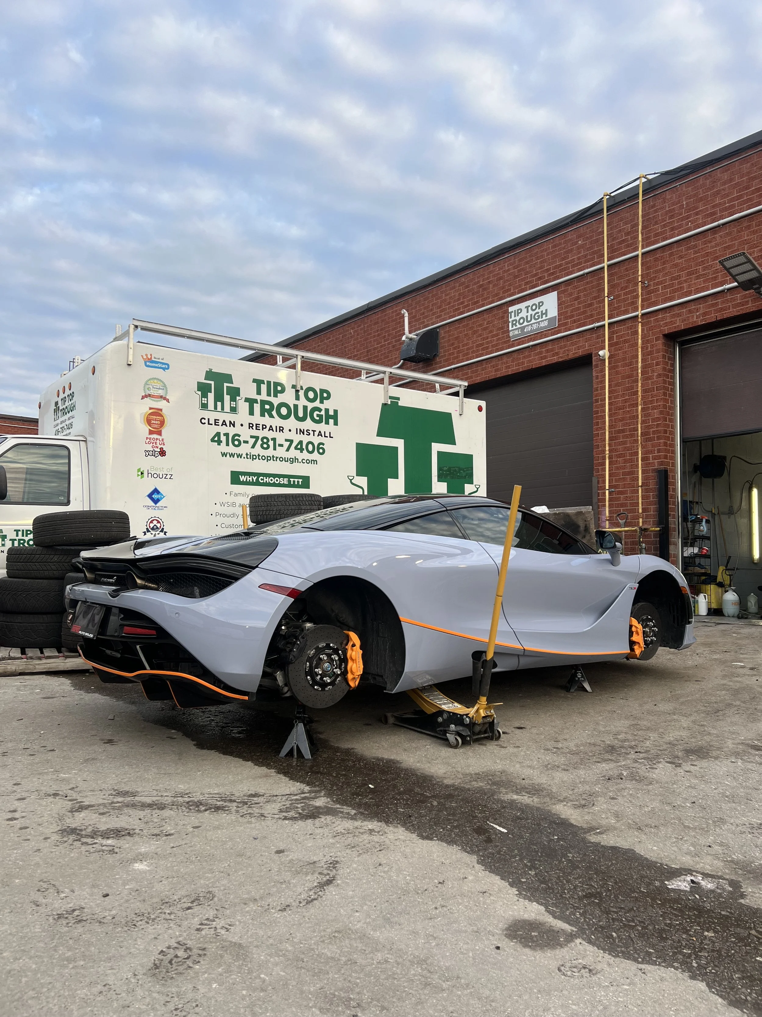 A silver sports car is on a lift outside an auto repair shop, missing its wheels and with Brake calipers painted orange. A white van with green branding and phone number is in the background.