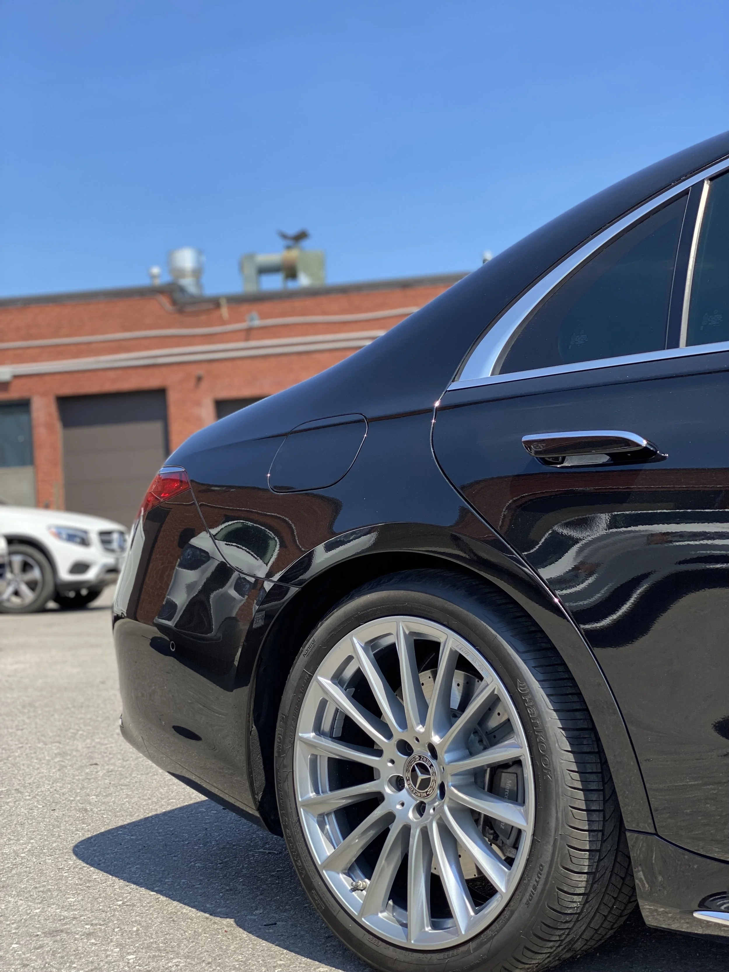 Close-up of the rear side of a black luxury sedan with silver alloy wheels, parked on a sunny street with a brick building and other cars in the background.