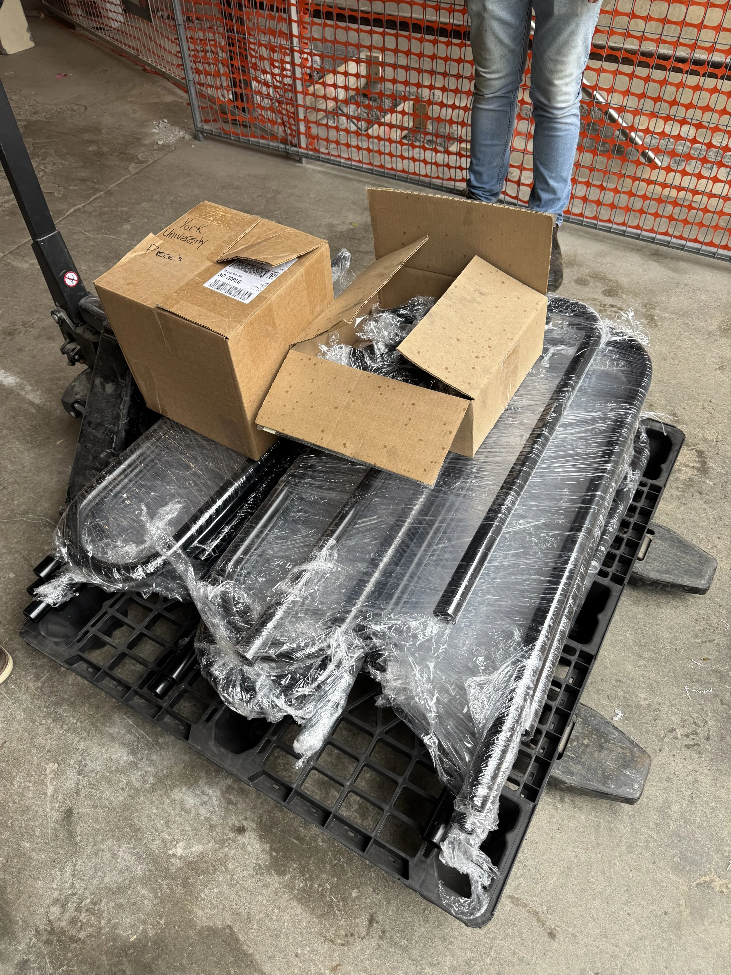 Unassembled black metal furniture parts wrapped in plastic on a black pallet, with two open cardboard boxes and a closed box labeled 'York University Pieces' on top, on a concrete floor with a red-orange plastic fence in the background