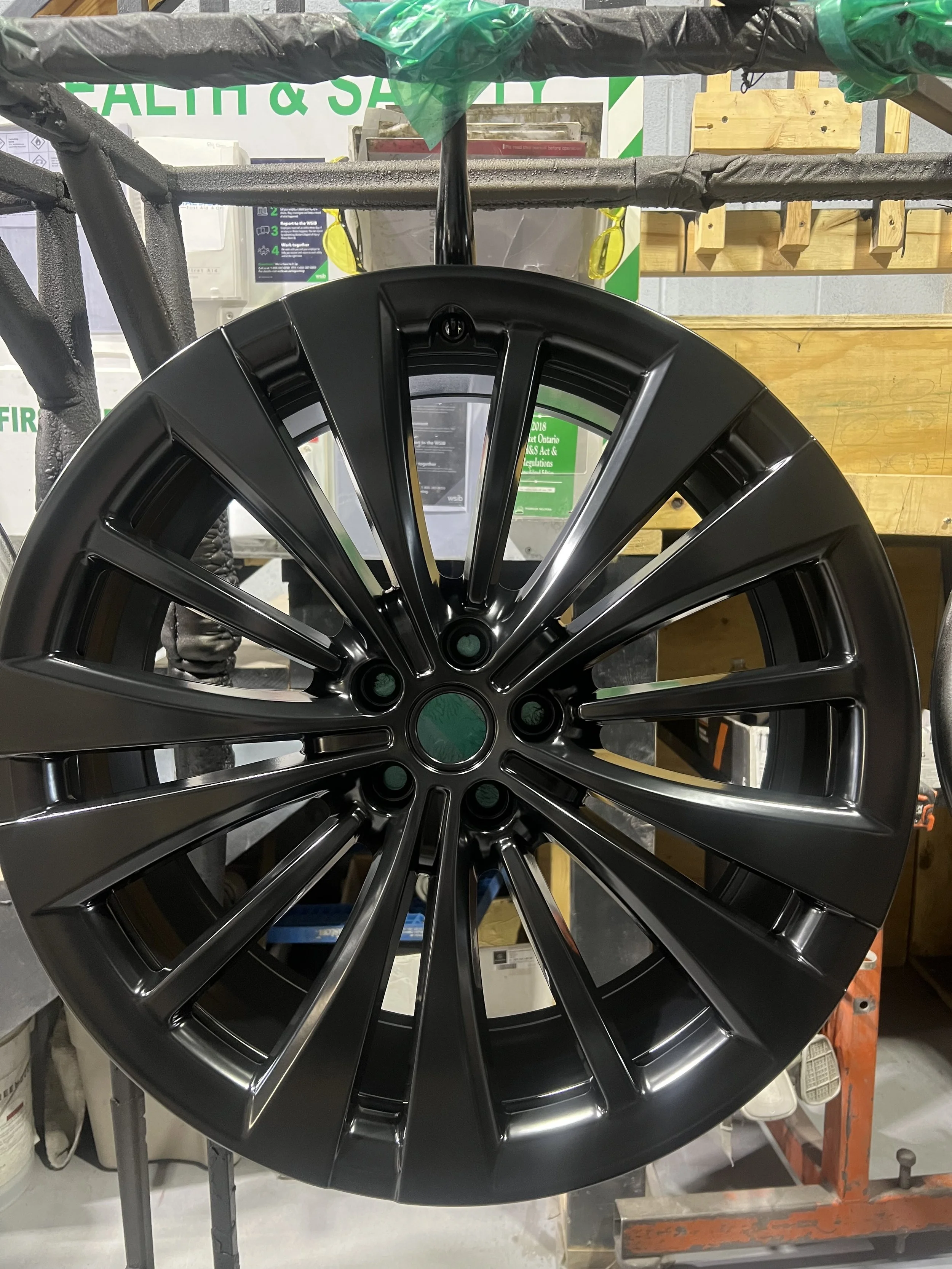 A black alloy wheel rim placed on a display stand in a workshop or store, with shelves and tools visible in the background.