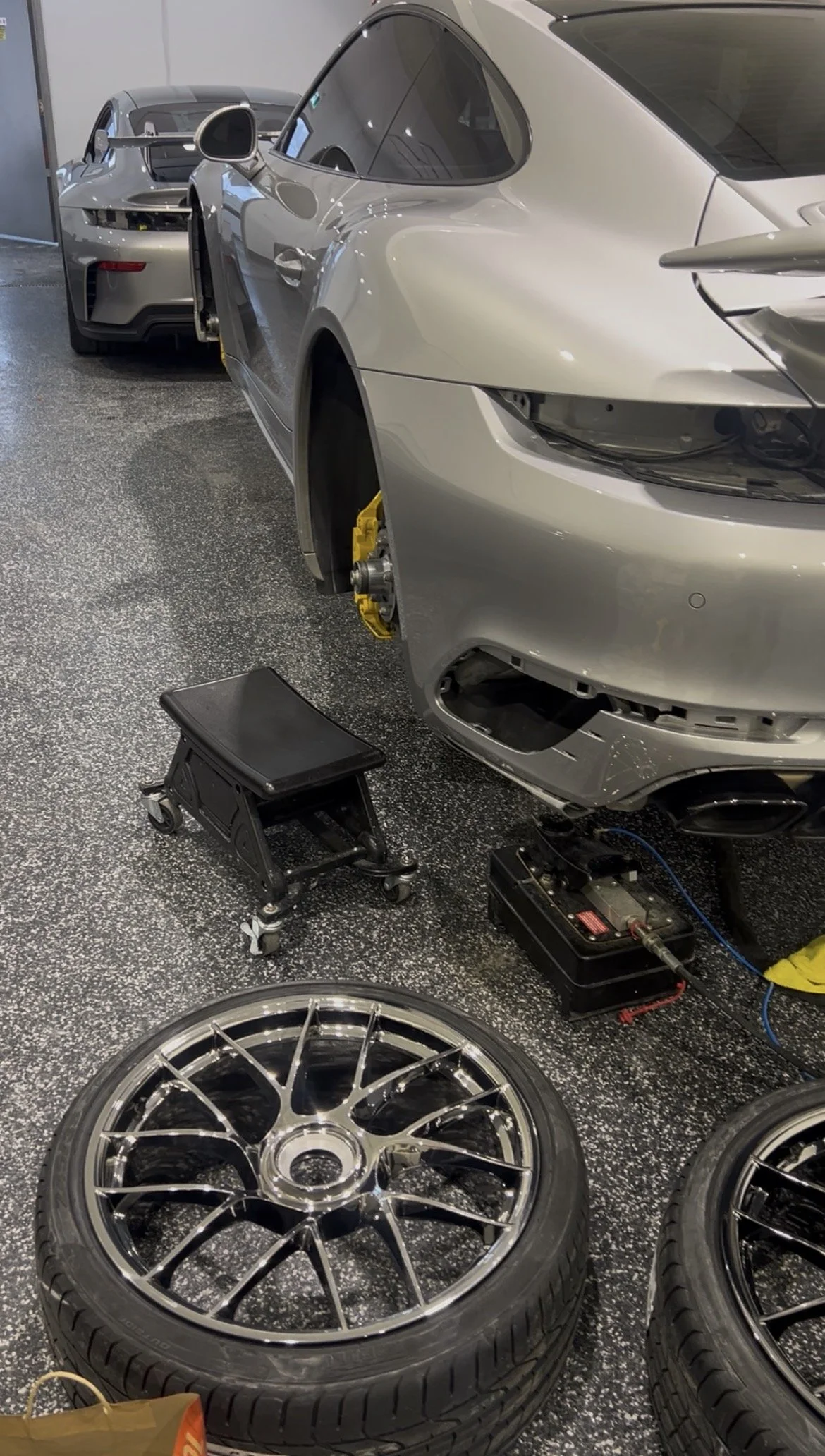 Two silver sports cars in a garage, with the front wheel of one car removed, showing brake tools and equipment, including a wheel, a stool, and diagnostic devices.