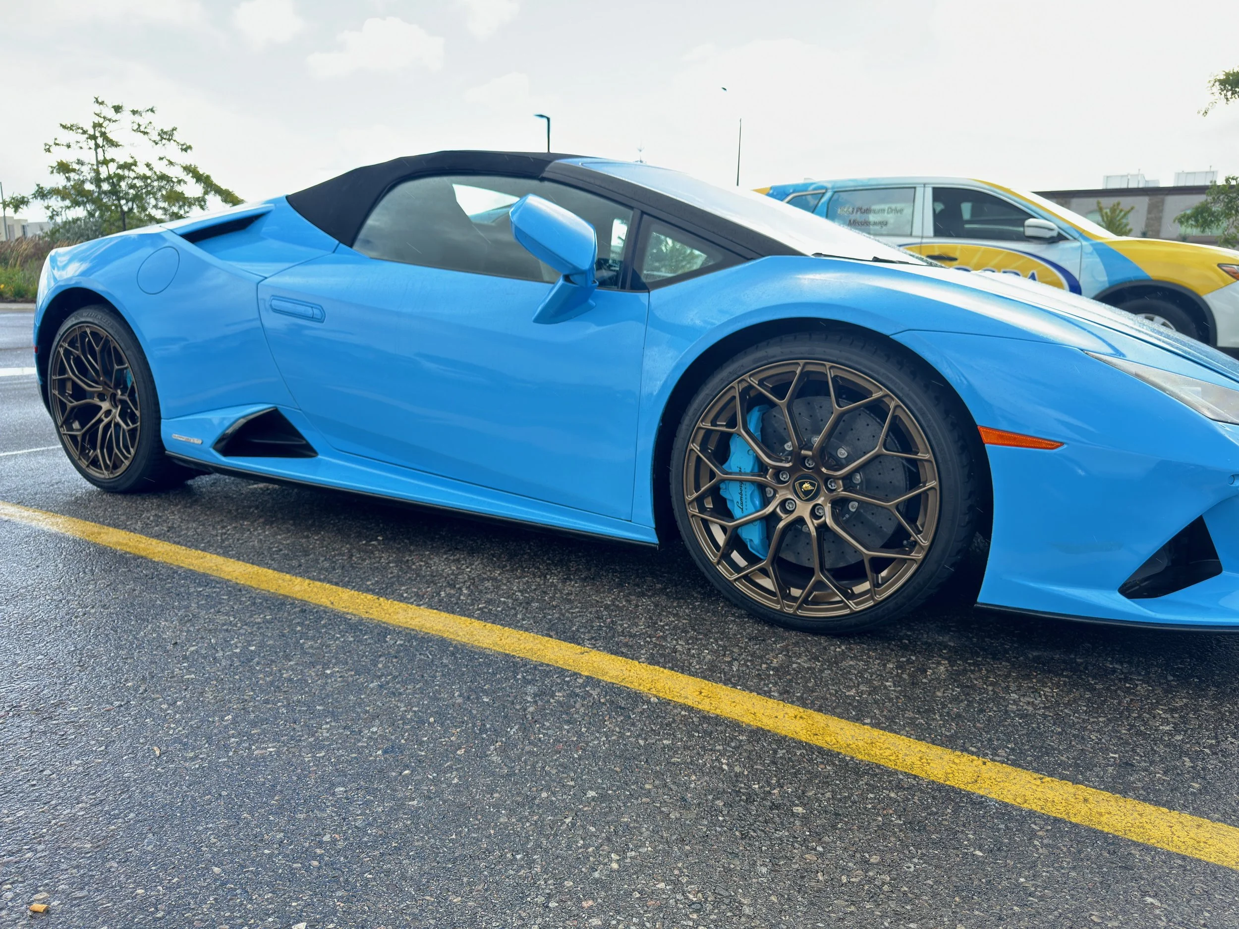 Blue Lamborghini Huracan convertible with dark wheels and gold brake calipers parked in a lot.