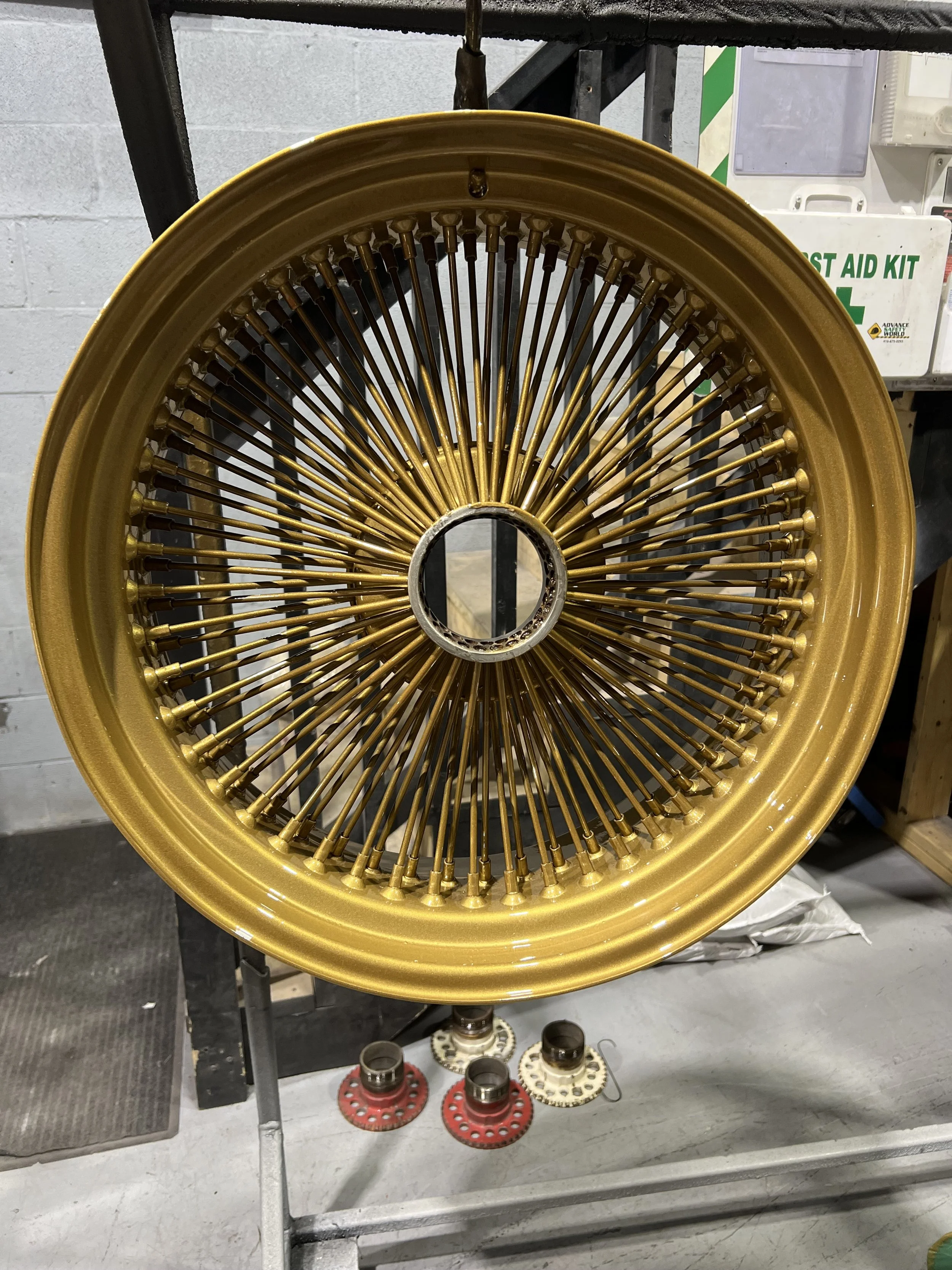 A gold wire-spoked wheel rim, likely for a vintage car, is mounted on a stand in a workshop setting. Some wheel parts and tools are visible at the bottom.