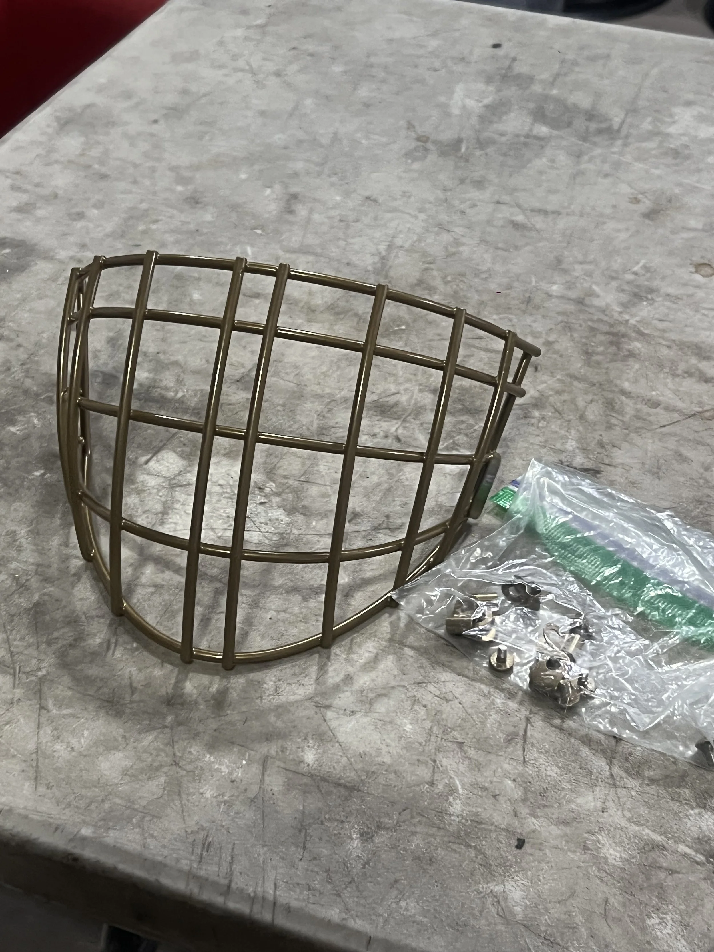 A metal cage with a curved shape, placed on a gray surface next to a plastic bag containing various small metal parts and screws.