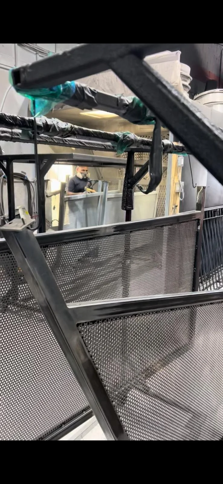 Metal framework and mesh panels in a workshop, with a person wearing a face mask working in the background.