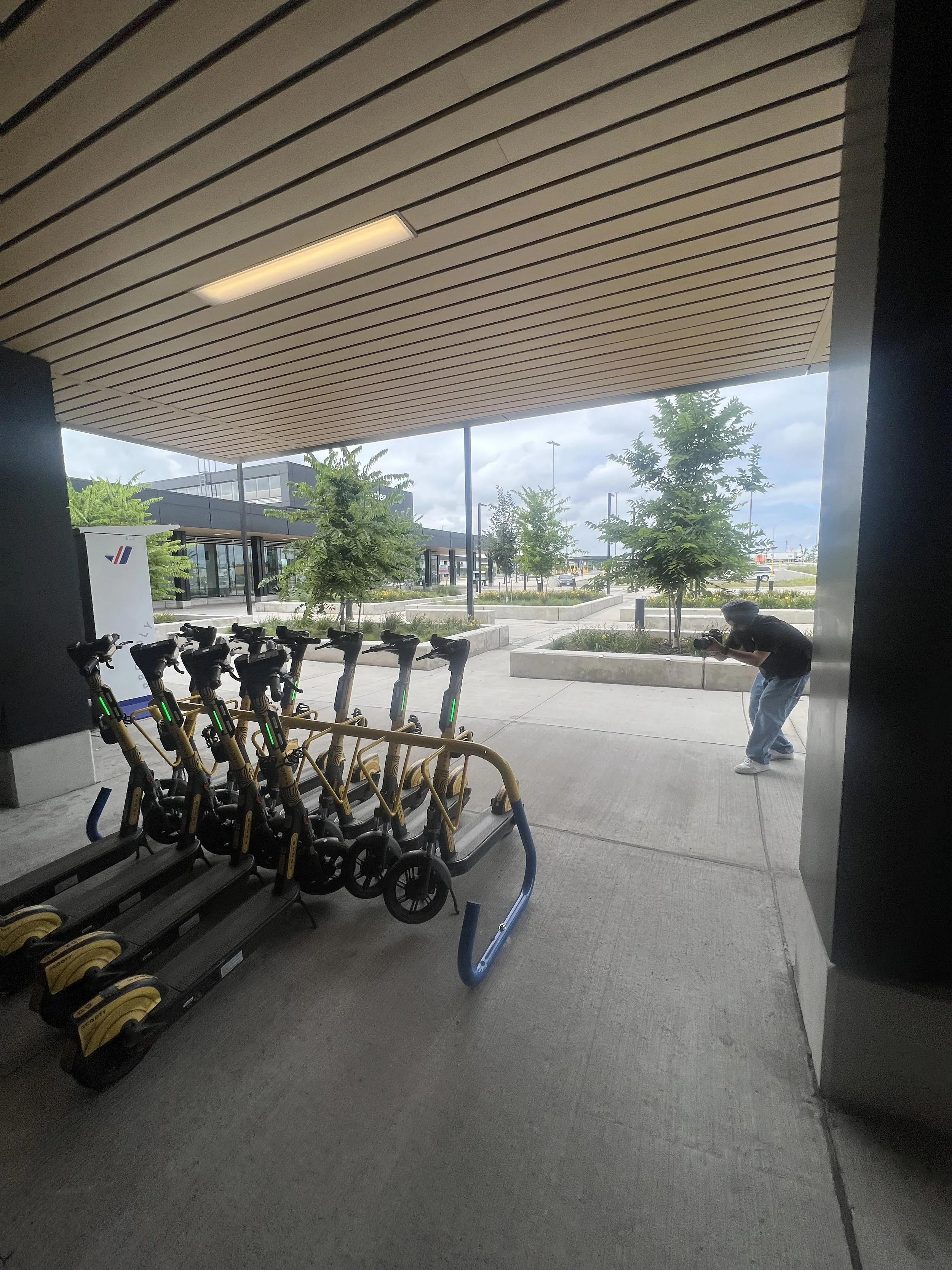 Electric scooters parked at a rack outside a building, with a person taking a photo in the background, greenery, and a cloudy sky.