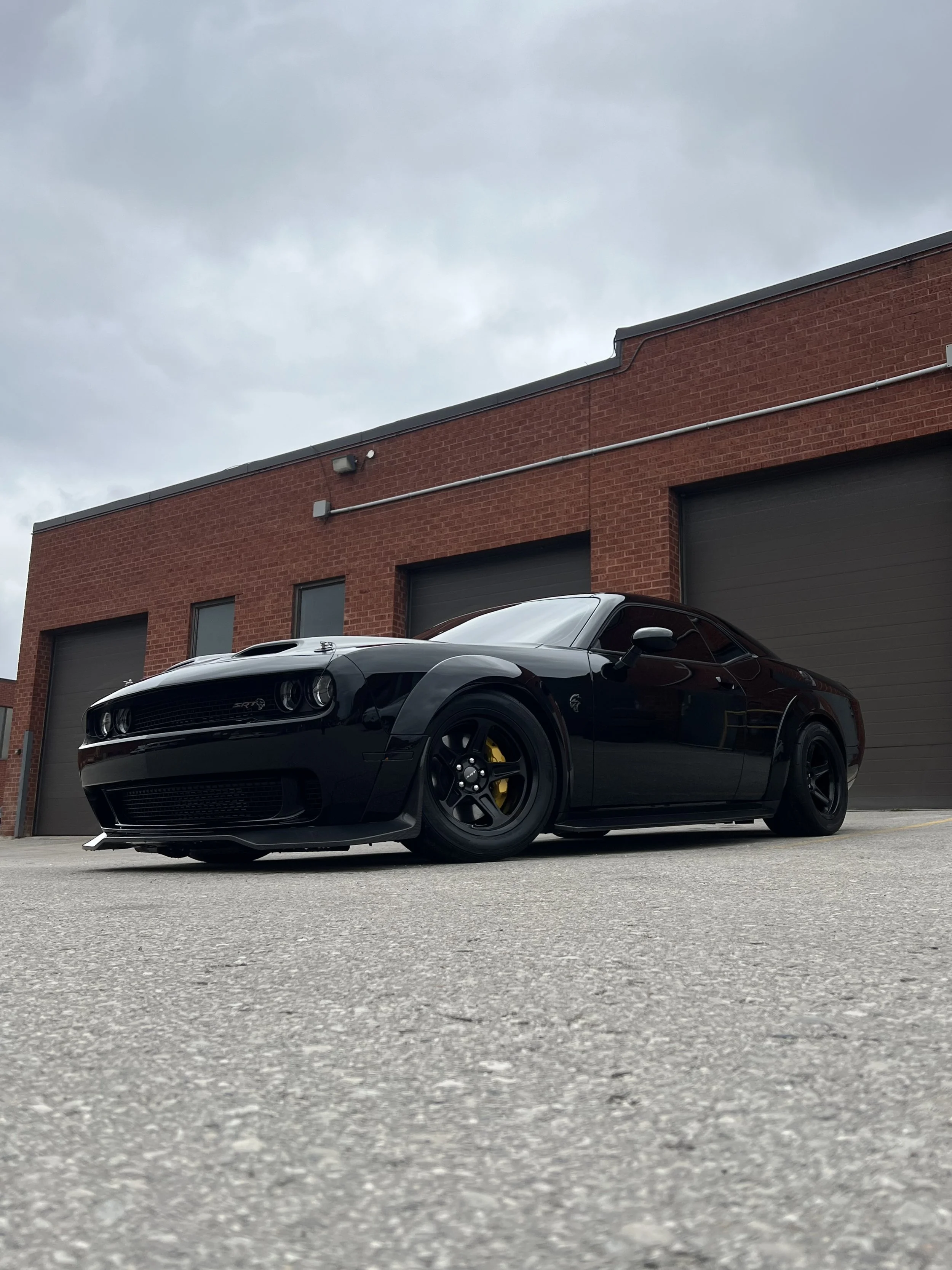 Black Dodge Challenger with yellow brake calipers parked outside a brick building with three garage doors on a cloudy day.