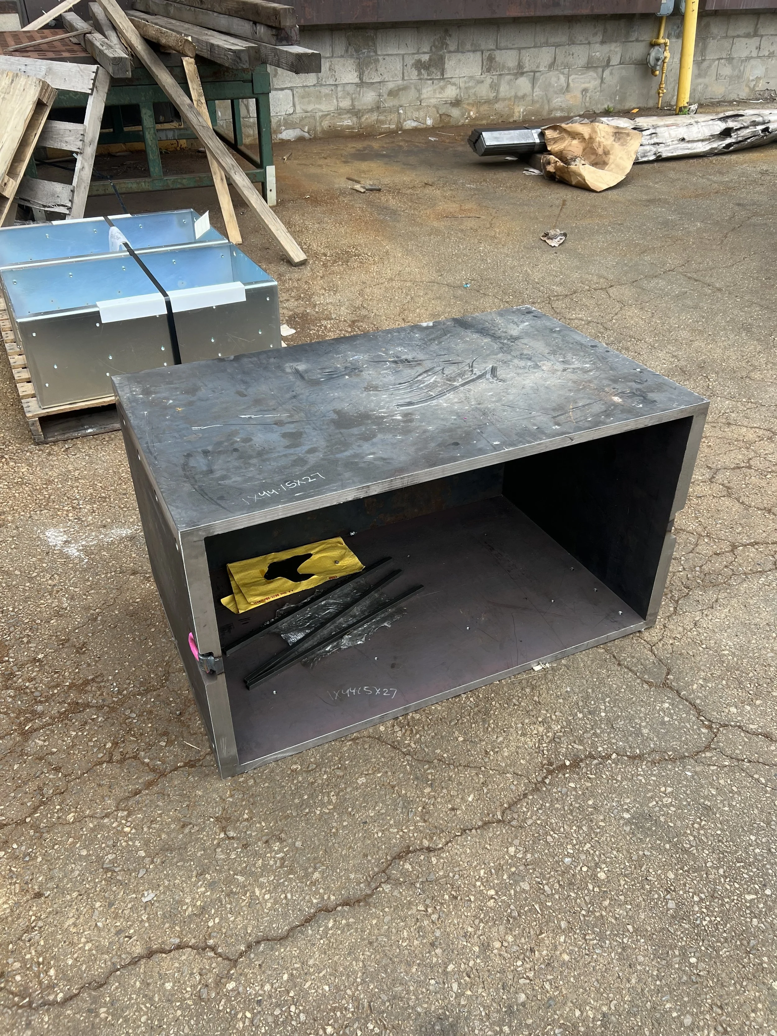 A metal table or workstation outdoors on cracked pavement with some tools and materials underneath, and a blue metal container in the background.