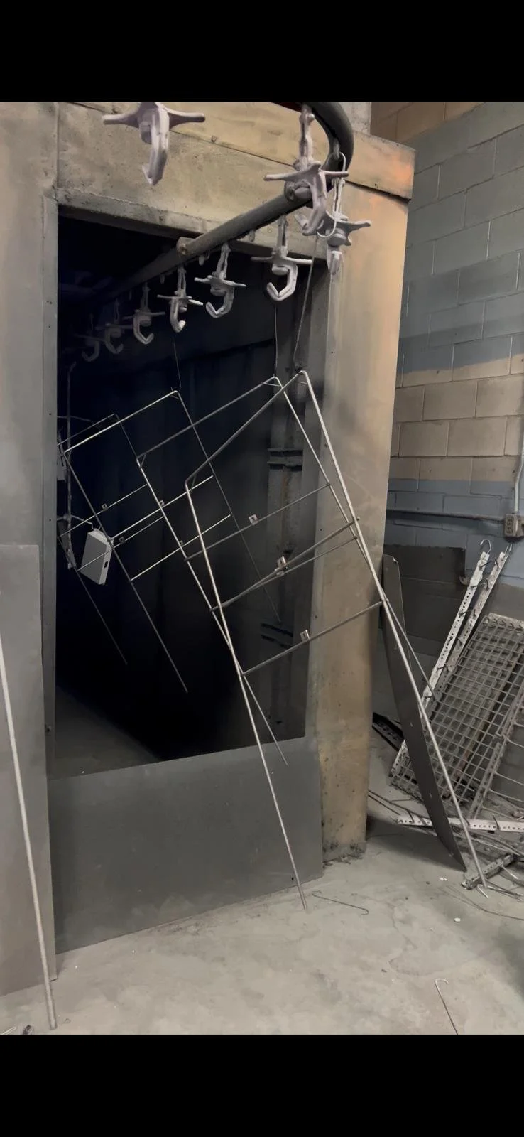A black metal tunnel or elevator shaft with metal hooks hanging from a pipe at the top. Several metal wire racks and a metal support are leaning against or inside the opening, with some debris and a pegboard on the floor nearby.