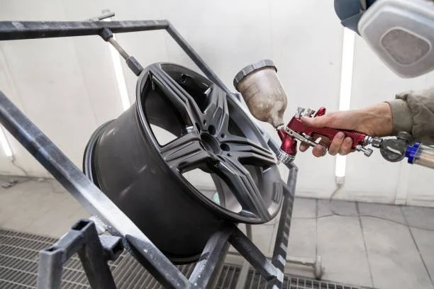 A person using a spray gun to paint a black alloy wheel in a workshop.
