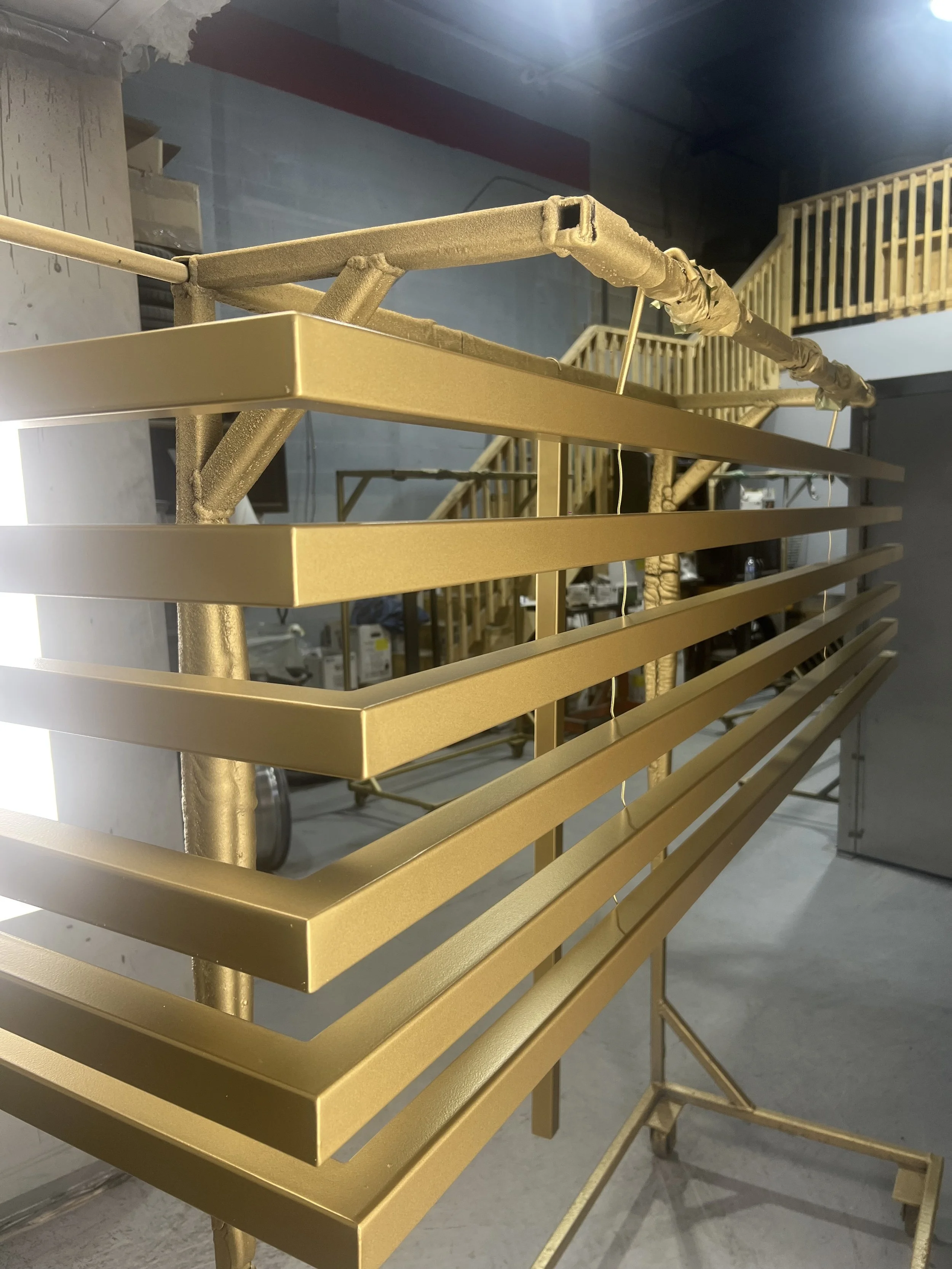 Metal racks in gold color, possibly part of a display or storage system, with a blurred background showing a workshop or industrial space.
