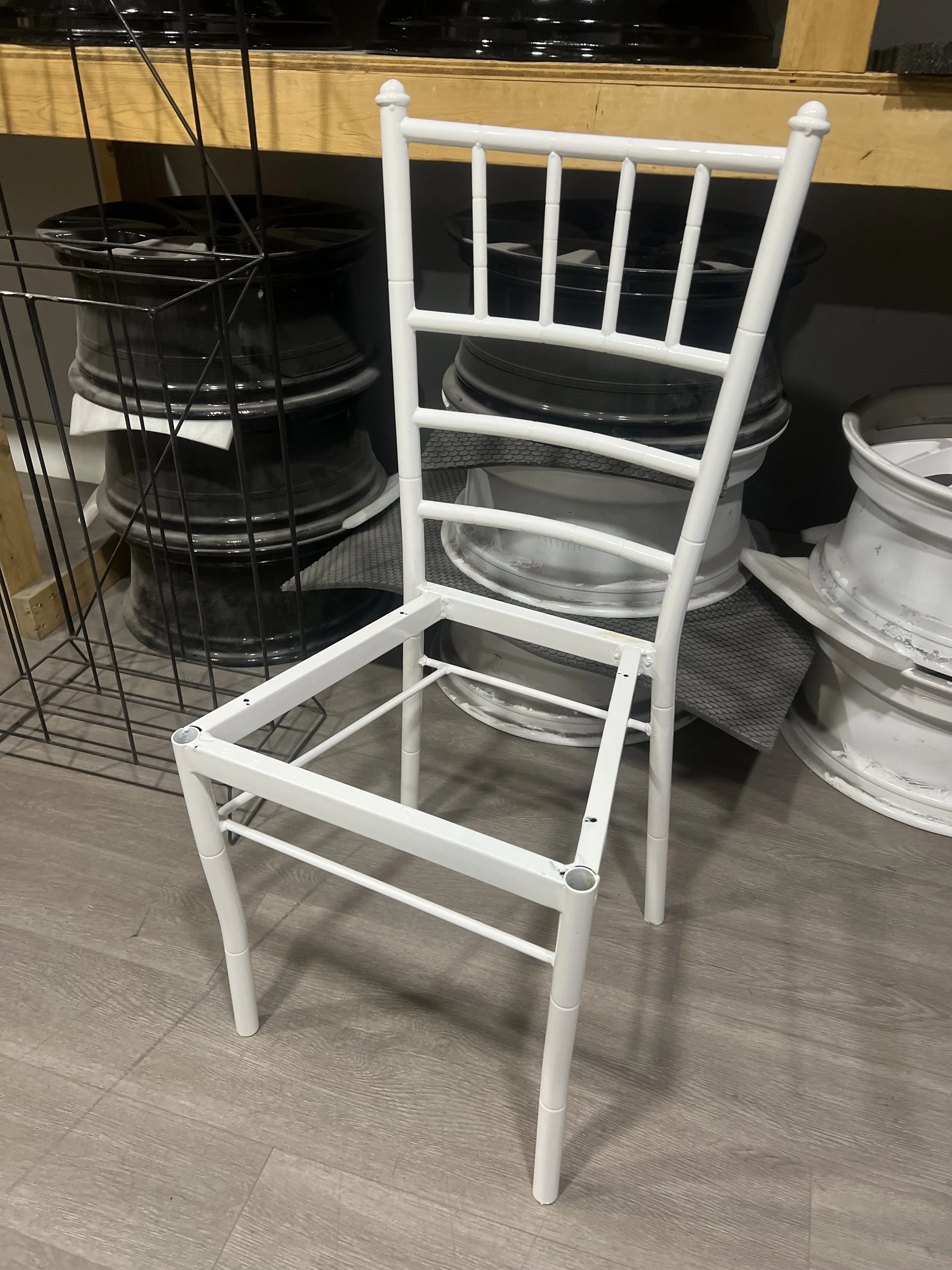 A white wooden chair with a slatted backrest and four legs, missing a seat, standing on a wood floor in front of a display of black and white wheel rims or trays.