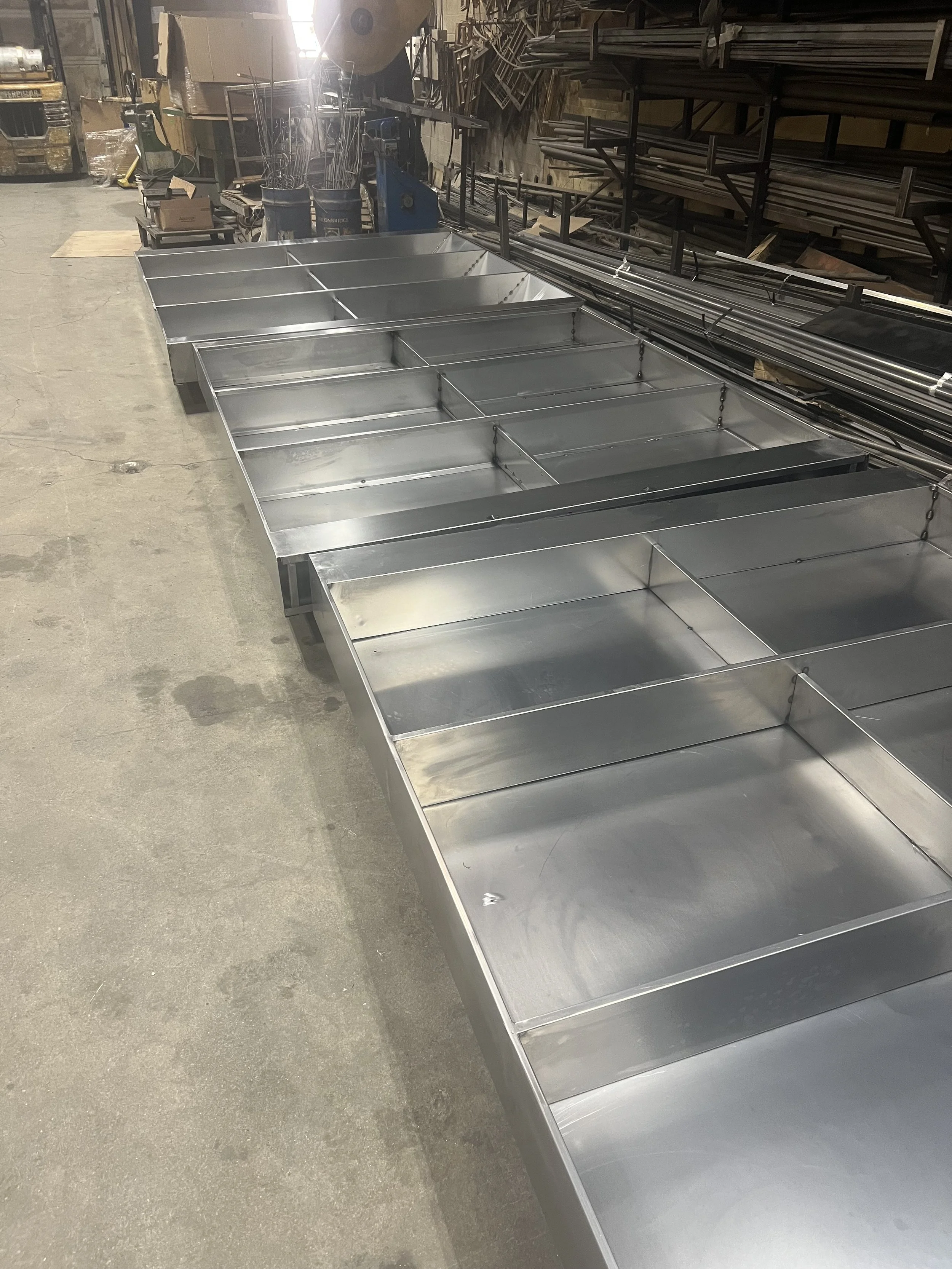 Several empty stainless steel tables or display cases in an industrial workspace, with various tools and materials on shelves and in the background.