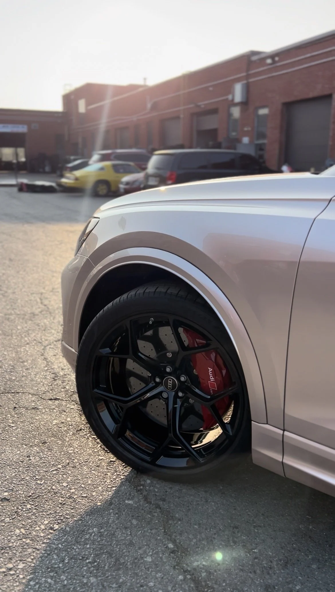 Close-up of the front right wheel of a silver Audi car, featuring a black rim and red brake caliper, parked on a gravel surface with a parking lot and brick building in the background.