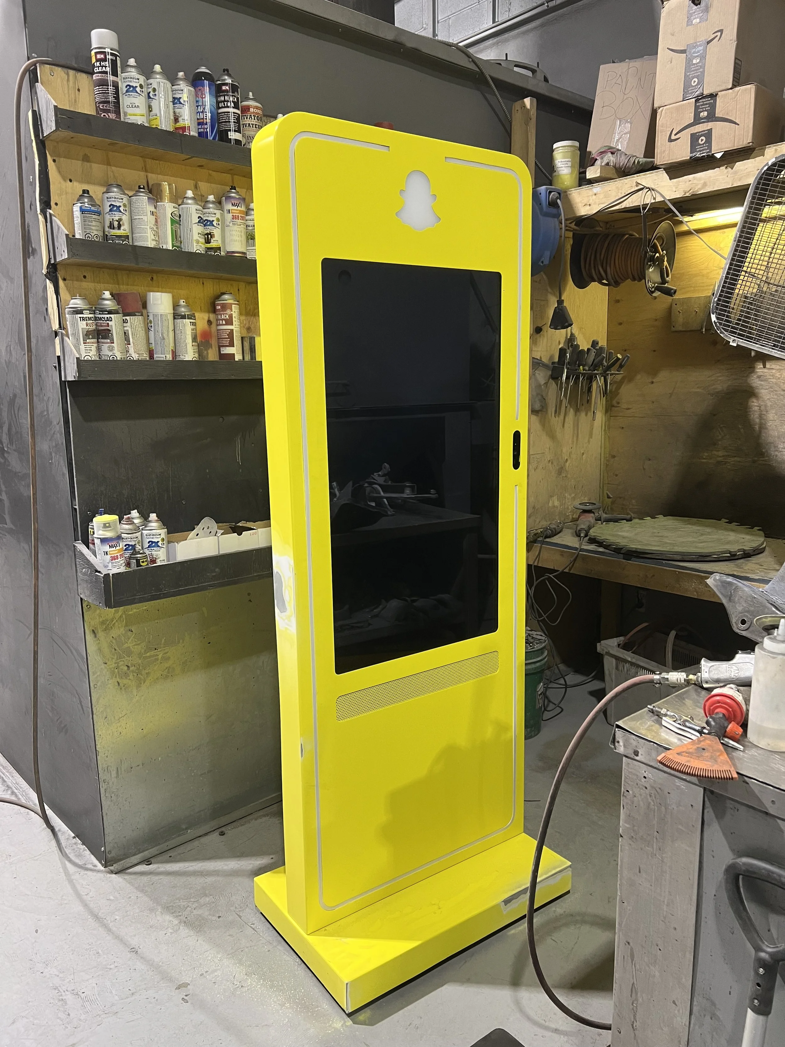 A bright yellow digital advertising kiosk with a black screen in a workshop or garage setting.